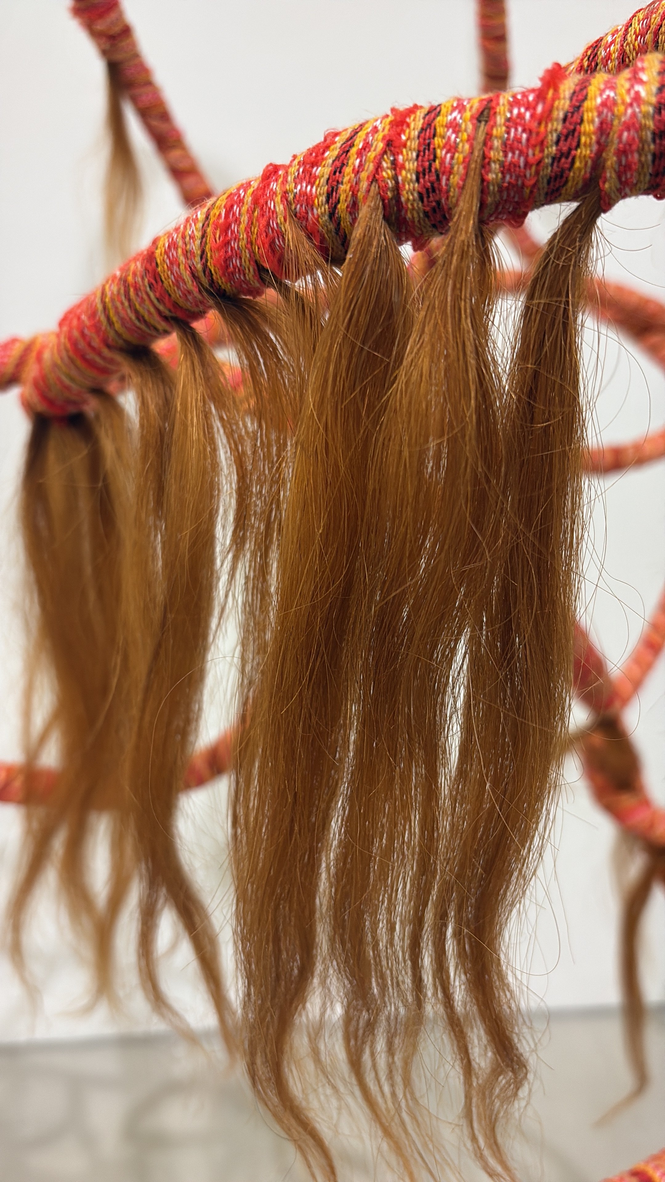 Close-up of a colorful, woven, hoop-shaped wall hanging with long, flowing, reddish-brown hair extending from it.