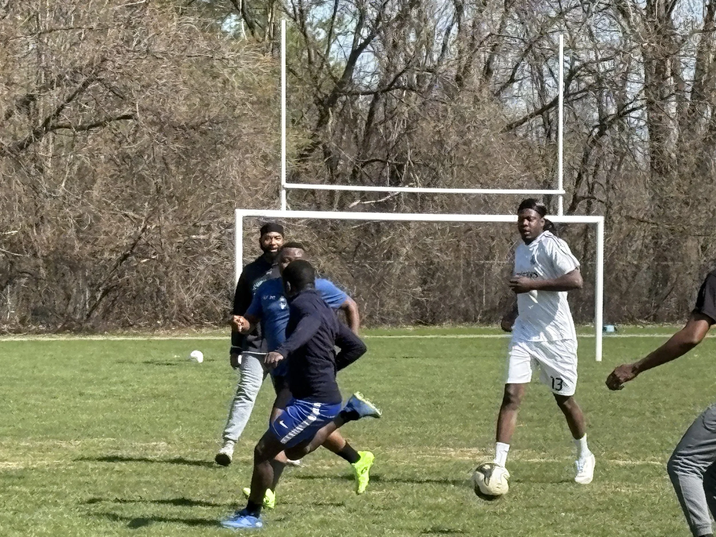 A group of people playing rugby on a green field with goalposts, surrounded by trees, under a clear sky.