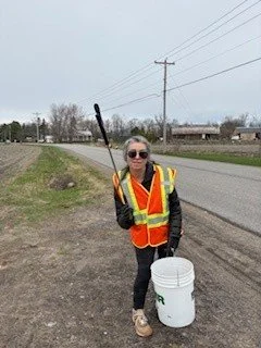 A woman wearing sunglasses and a reflective safety vest holding a trash picker and a white bucket, standing on a grassy roadside near a paved road with power lines, trees, and houses in the background.