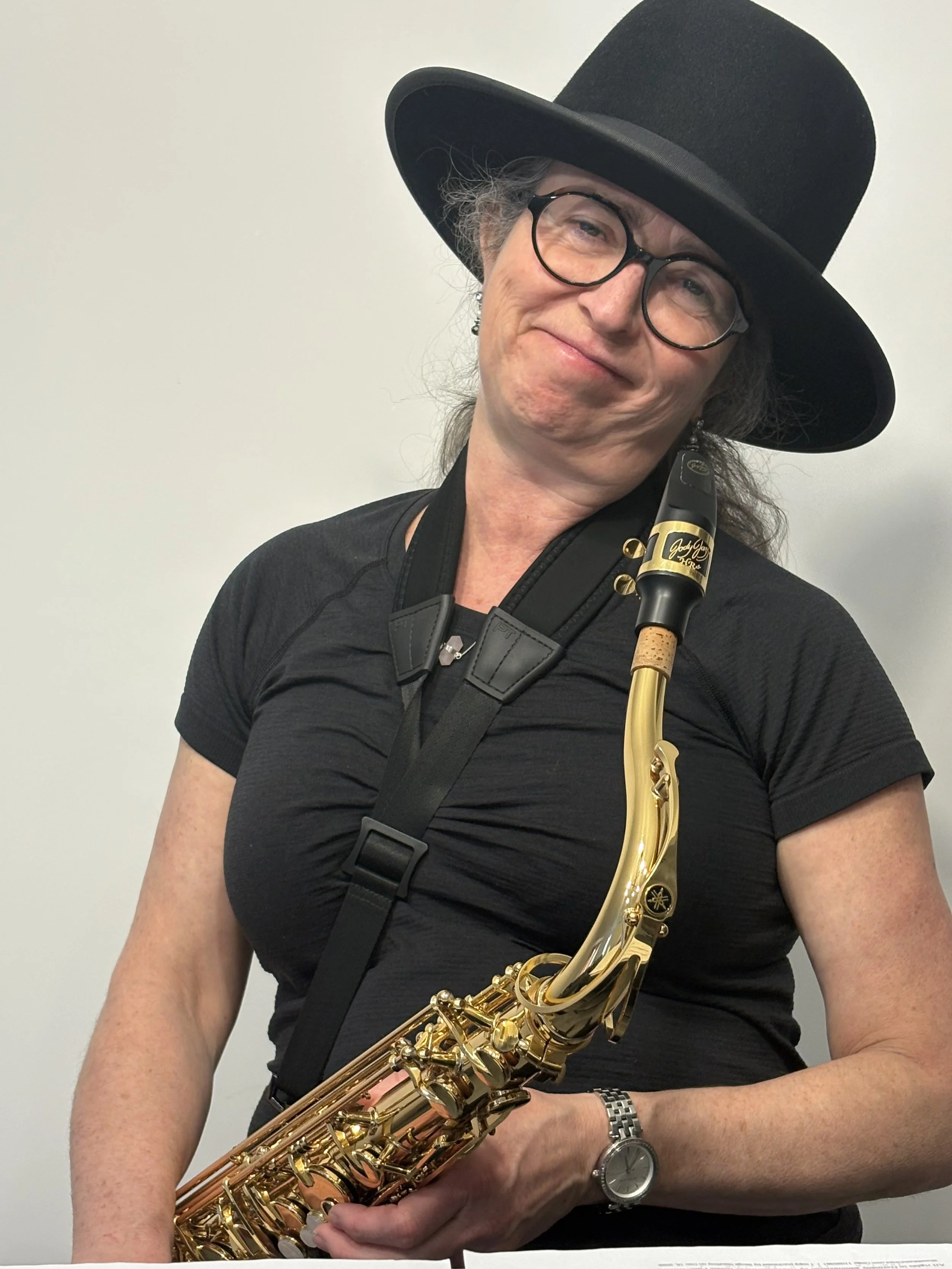 A person wearing a large black hat, black glasses, a black shirt, and a silver watch is holding a shiny gold saxophone. The background is plain and light-colored.