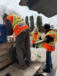 Three people wearing reflective safety vests and masks working outdoors, handling buckets on a wooden platform.