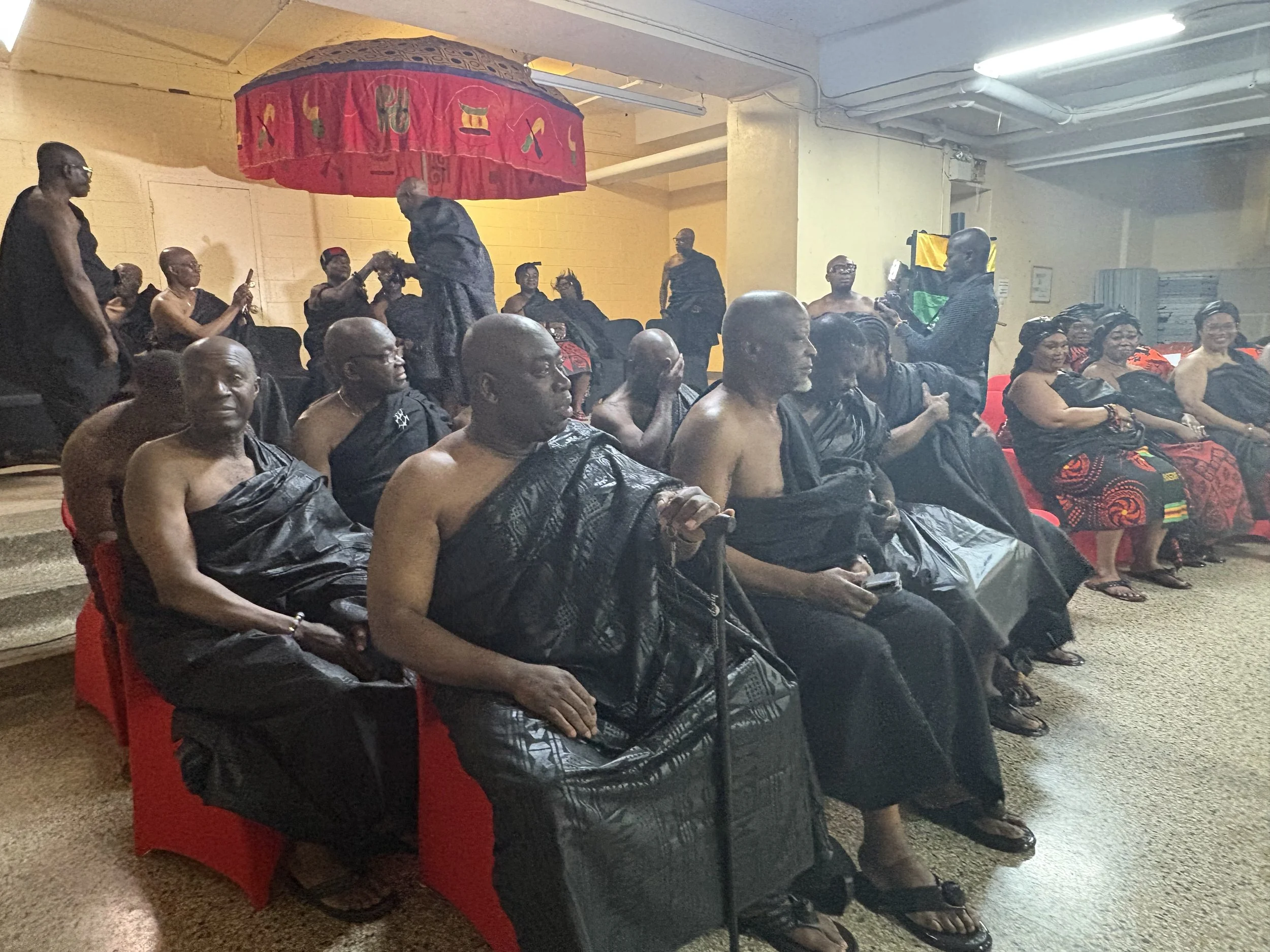A group of people dressed in traditional African attire and some shirtless, sitting and standing in a room, with a decorative red and black umbrella overhead. They appear to be participating in a cultural or ceremonial event.