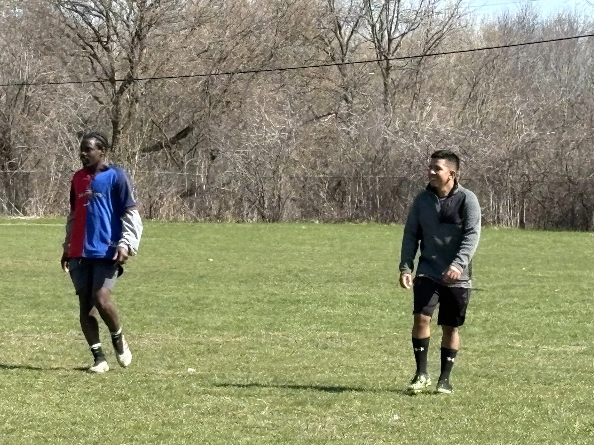 Two young men playing soccer on a grassy field with trees in the background, during daytime.