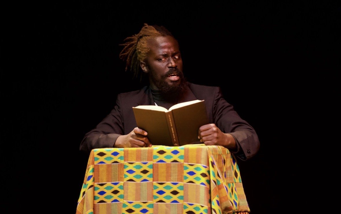 A man with dreadlocked hair and a beard reads a book while sitting at a table covered with a colorful African print cloth against a black background.