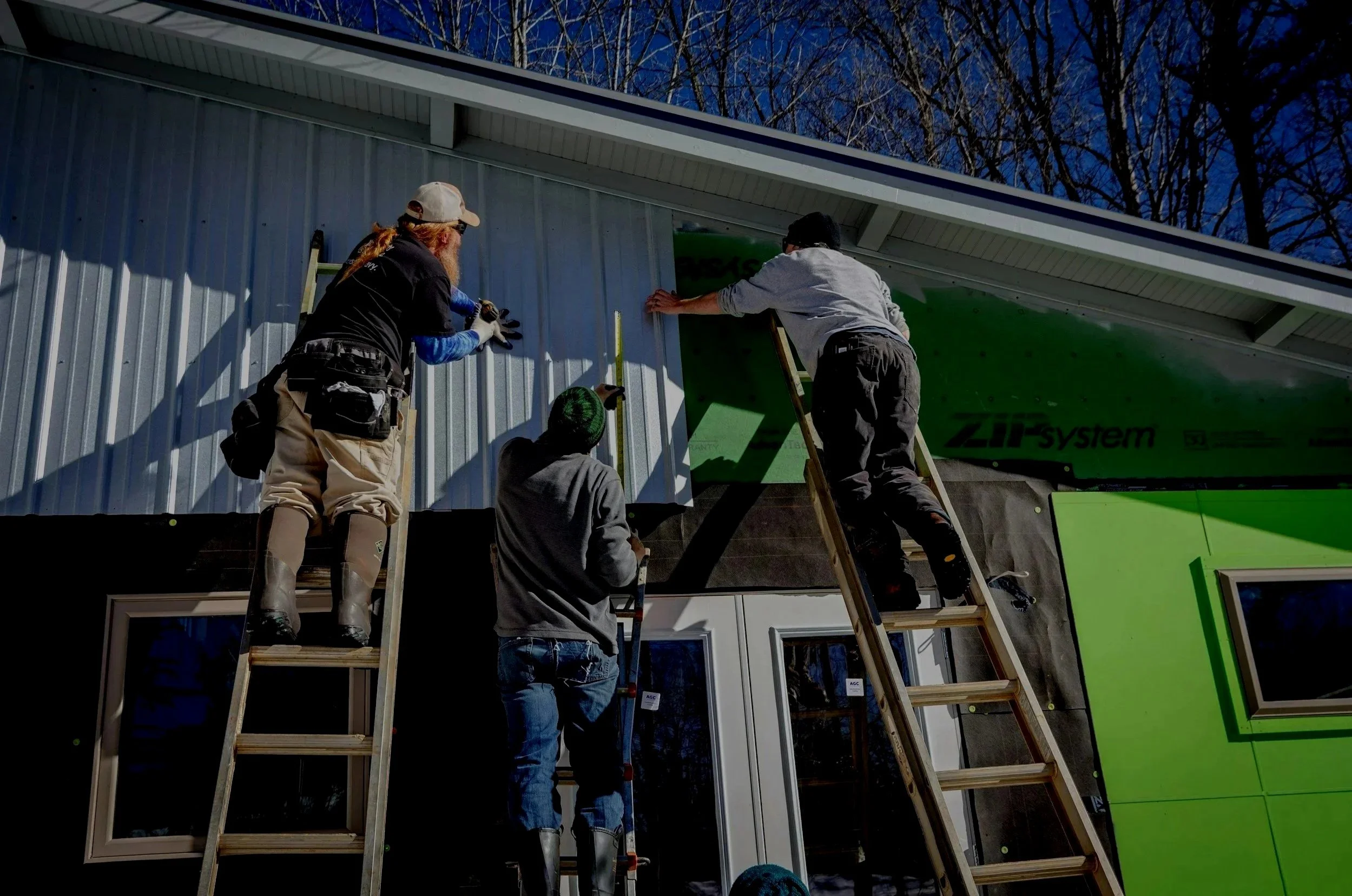 Four workers install green and metal siding on a modern building exterior. They use ladders to reach the upper part of the wall during daytime.