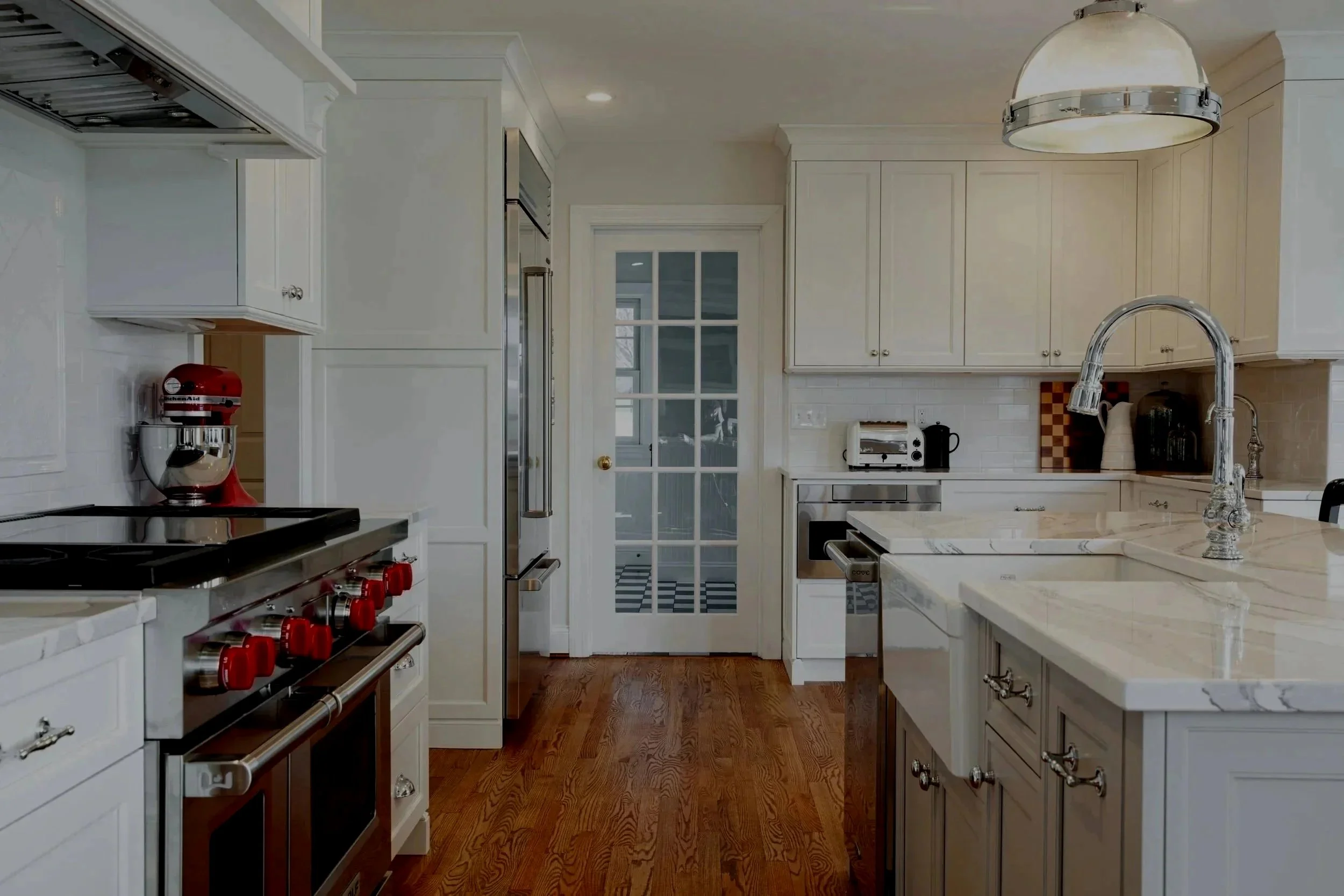Modern kitchen with white cabinets, marble countertops, wooden flooring, stainless steel appliances, a red stand mixer, and a glass-paneled door leading outside.