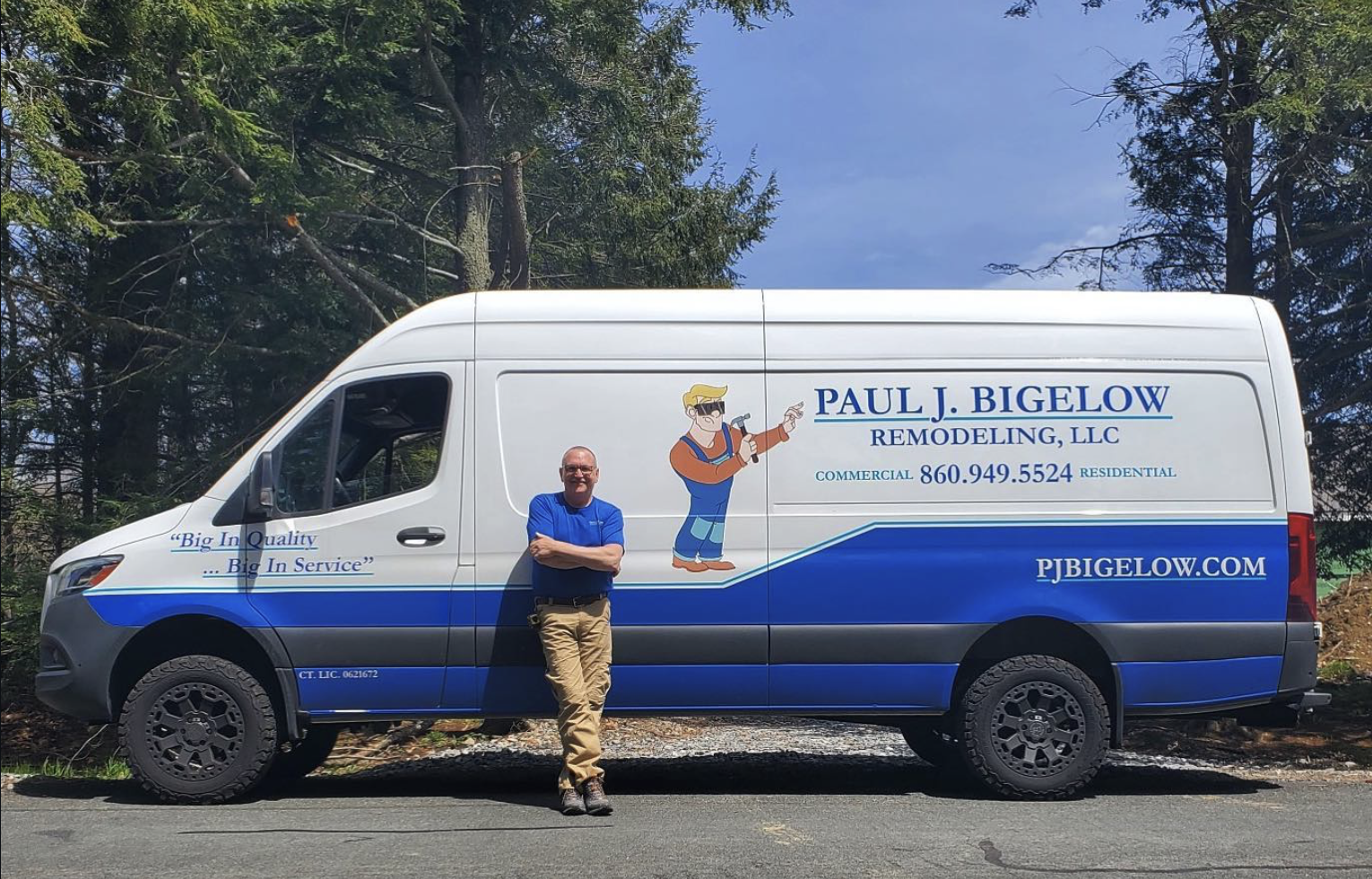 A man in a blue shirt and khaki pants standing in front of a white and blue remodeling service van with company information and cartoon of a handyman, parked on a road with trees in the background.