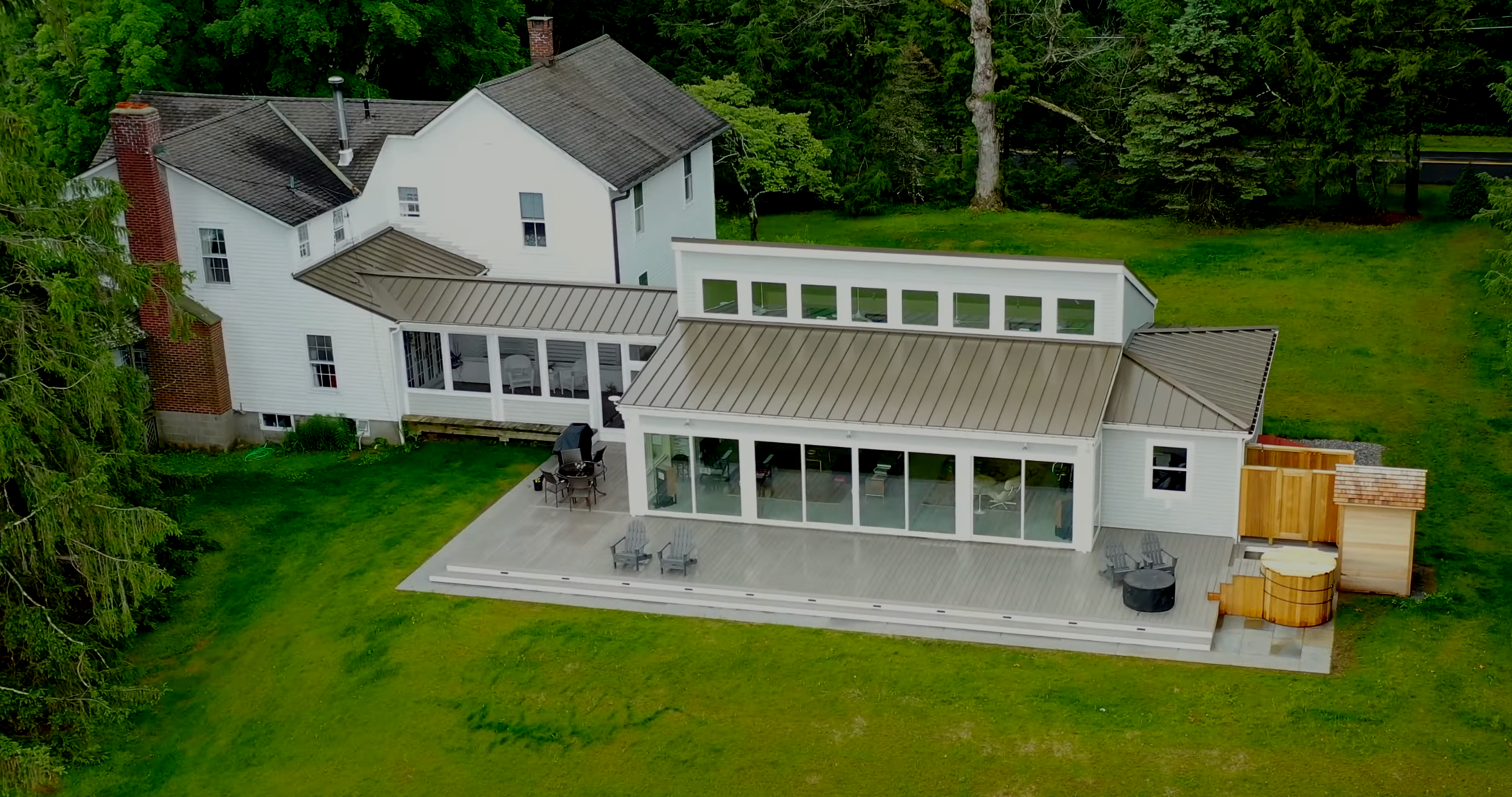 A large white house with multiple roof sections and a chimney, surrounded by green trees and lawn, with a spacious wooden deck in the backyard featuring outdoor furniture and a hot tub.