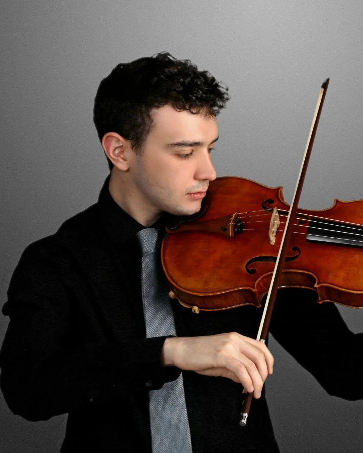 Young man with dark curly hair playing a violin, wearing a black suit and gray tie against a plain gray background.