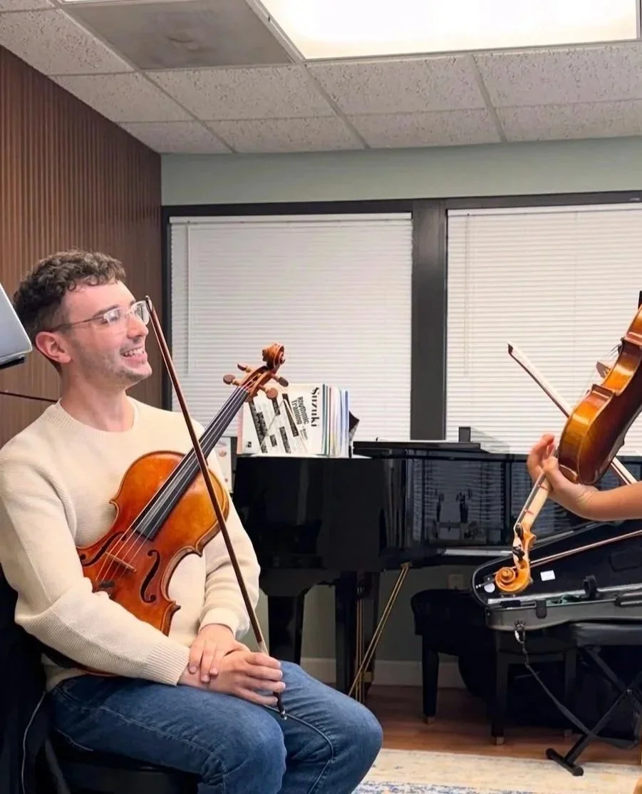 A man smiling while holding a violin sits in a room with a grand piano and sheet music, two people are likely rehearsing or playing music together.