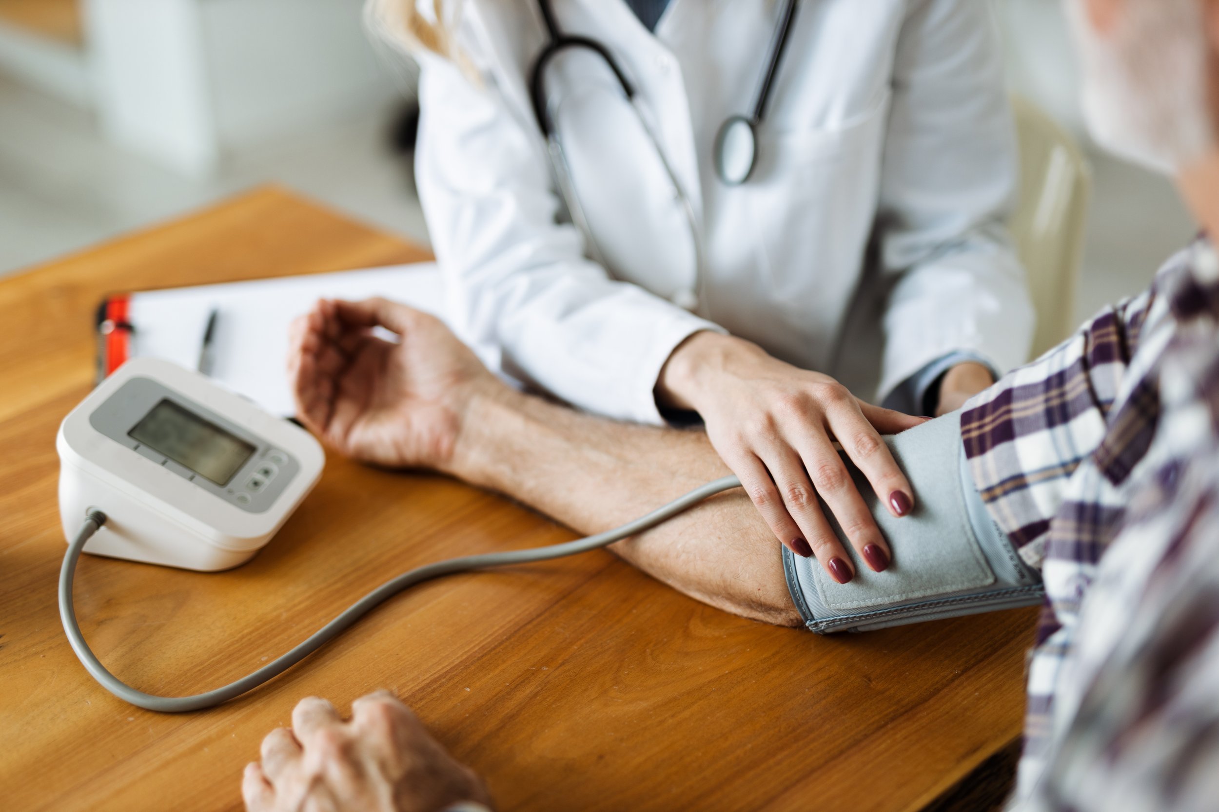 A healthcare professional taking a patient's blood pressure using a sphygmomanometer, with a digital monitor on a wooden table.