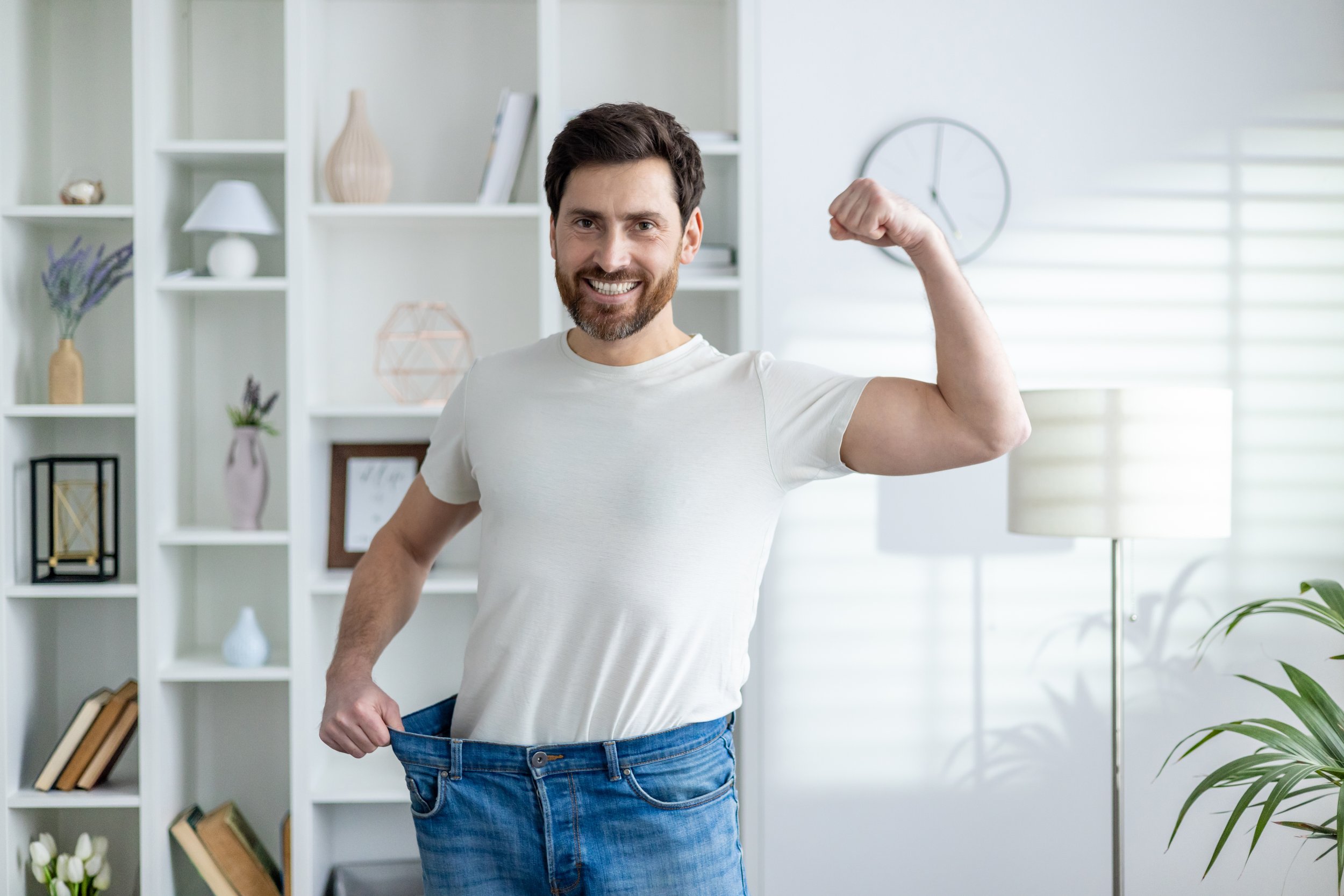 Man smiling and flexing arm muscle while holding out his empty jeans to show weight loss in bright living room.
