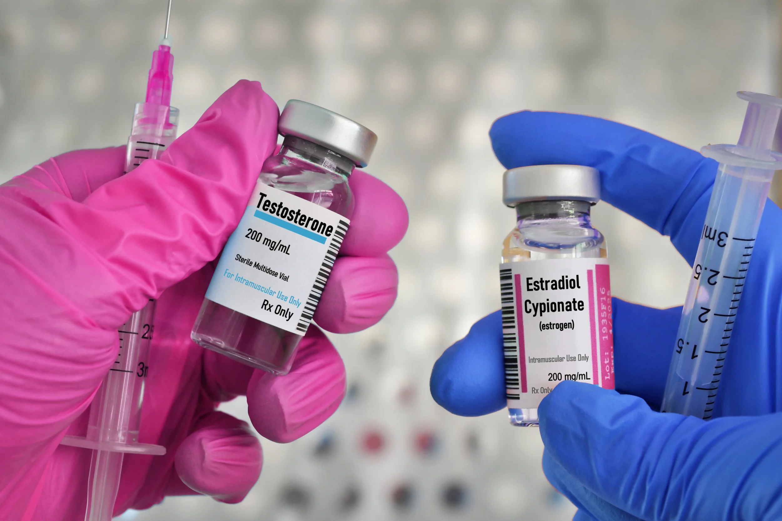 Hands in pink and blue gloves holding vials of testosterone and estrogen medications with syringes, set against a blurred background.