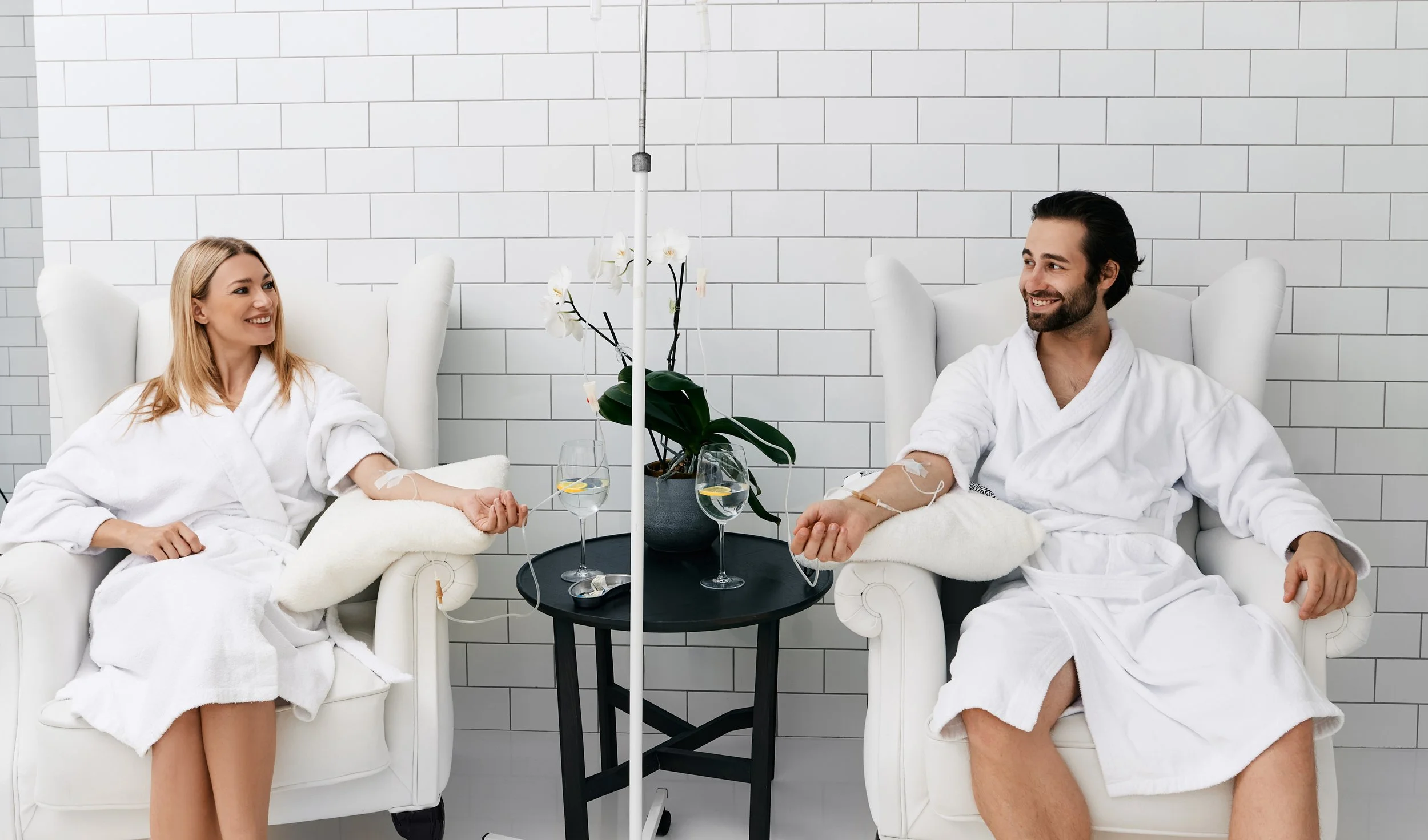 Two people in white robes sitting in white armchairs, donating blood through IVs, with a black table between them holding glasses of water and a plant, in a bright room with white tiled walls.