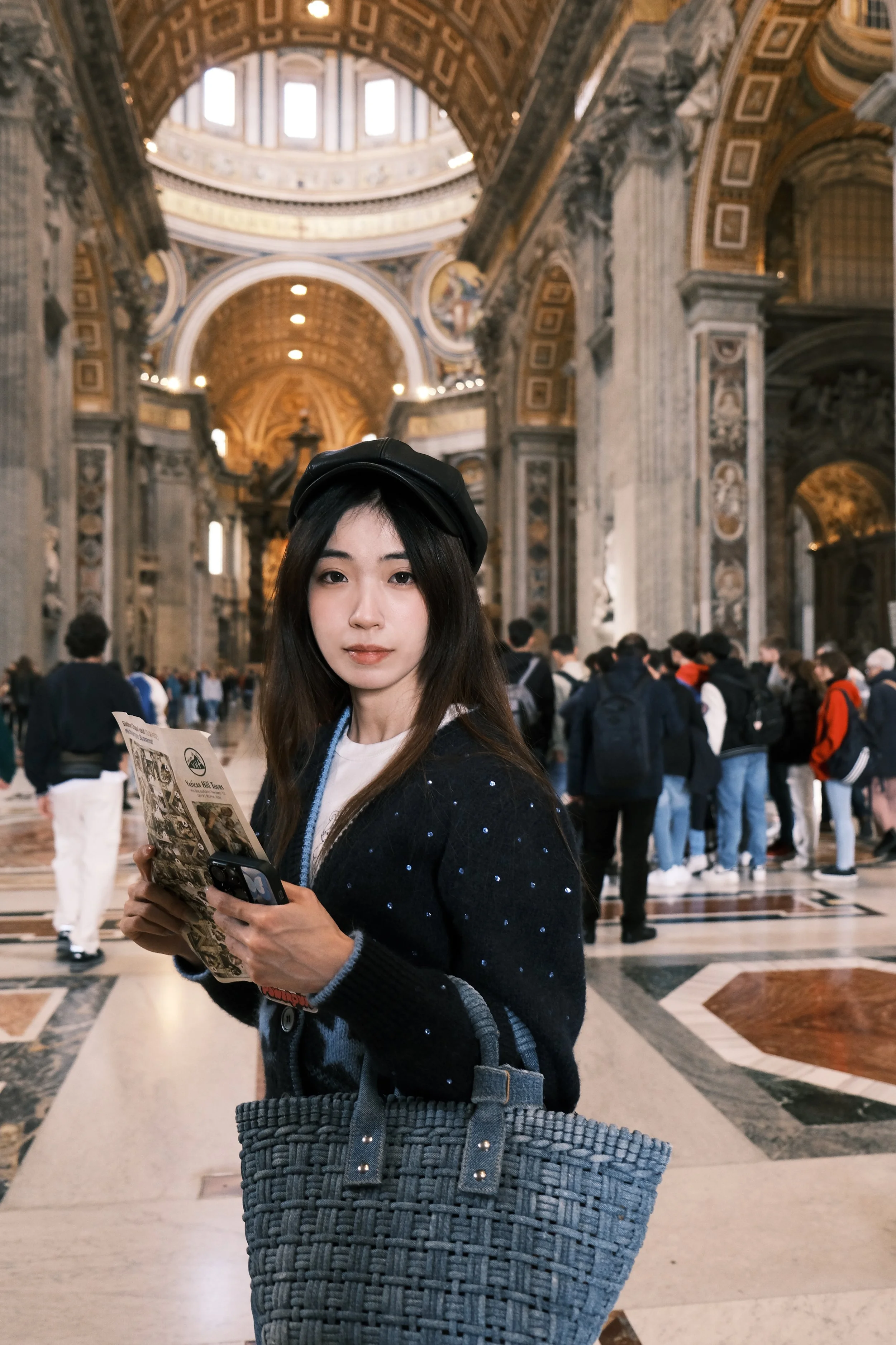 A young woman holding a brochure and smartphone, standing inside a grand cathedral with ornate architecture, high ceilings, and a crowd of visitors in the background.