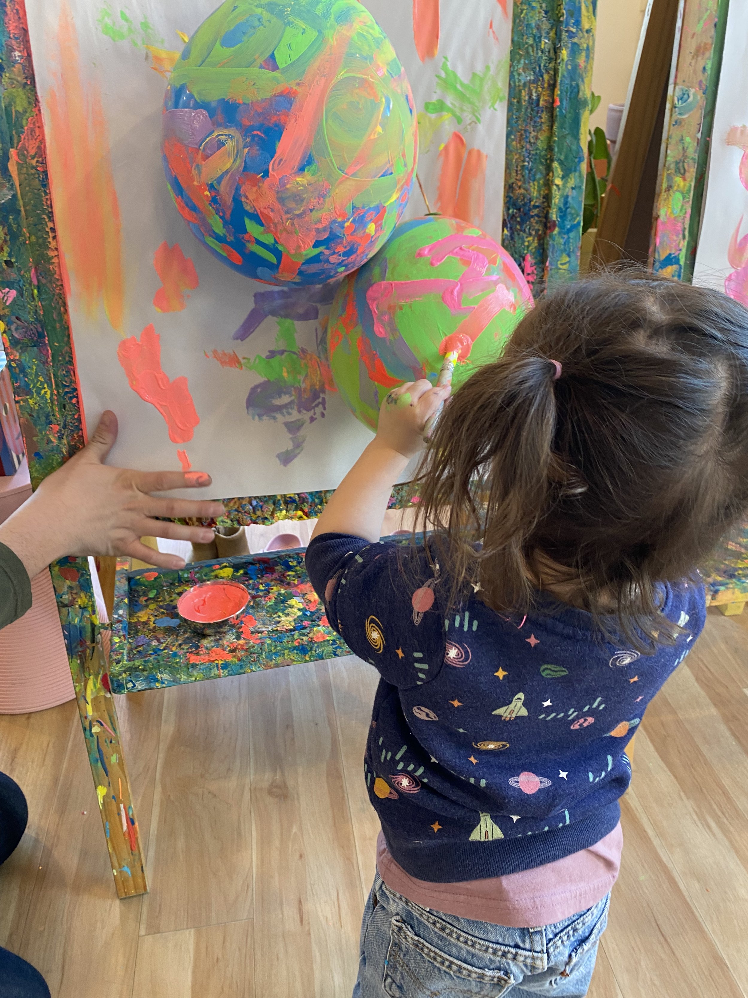 A young girl with brown hair tied back in a ponytail, wearing a navy blue shirt with galaxy and space themed prints, is painting on a large white sheet attached to a colorful, splattered table surface. She is painting pink and orange colors on two large, painted paper-mâché globes that are decorated with bright, abstract patterns, resembling planets.