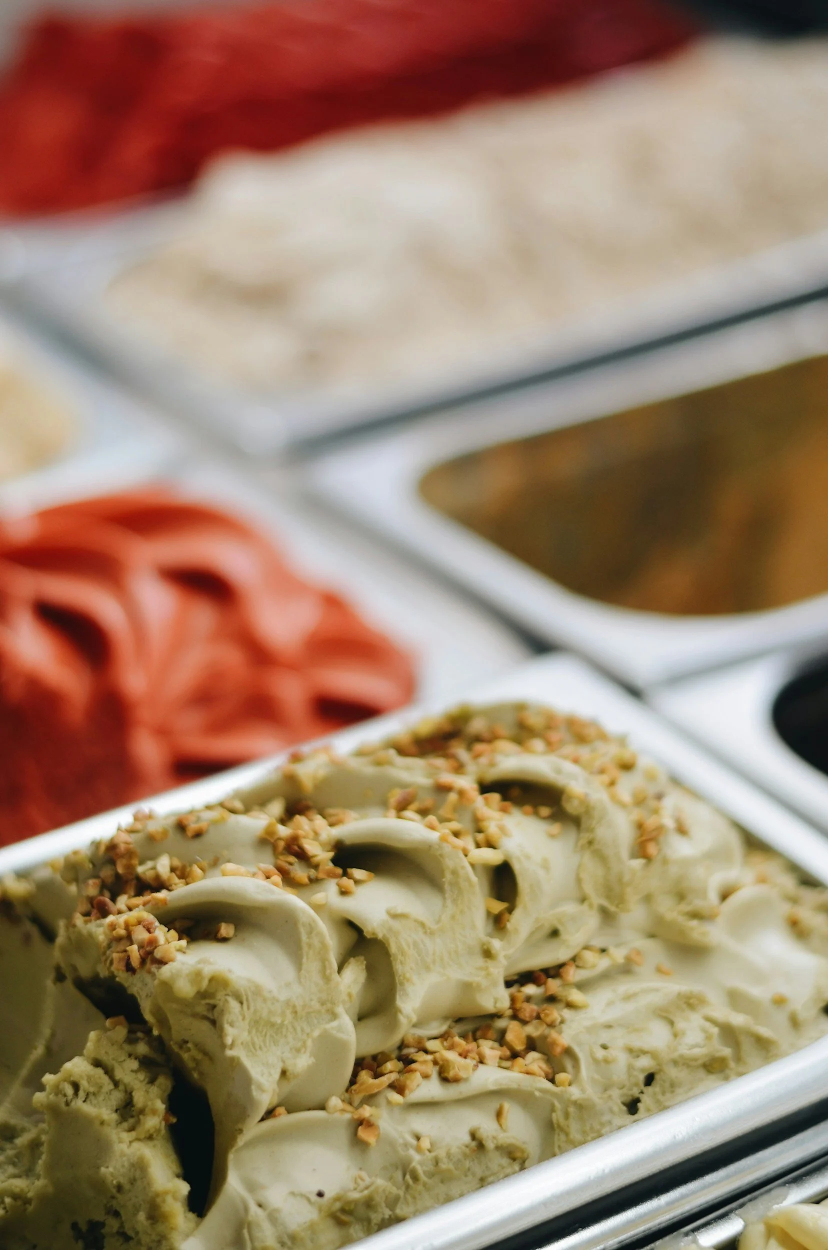 Tins of gelato on display in a gelato shop, in the forefront is a pale green pistachio gelato topped with chopped pistachios