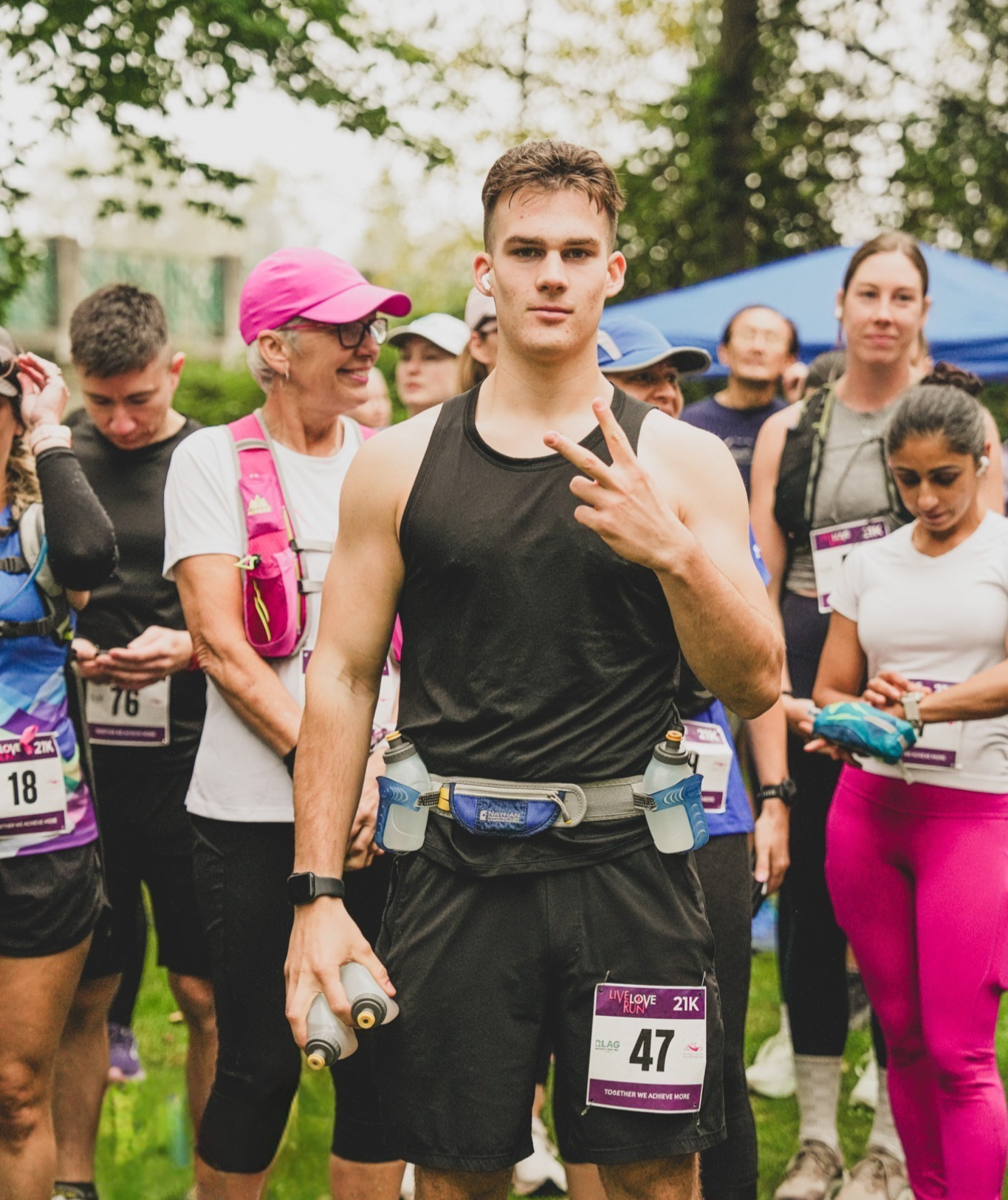 A young man holding a water bottle and making a peace sign at a group running event, with other runners and green trees in the background.