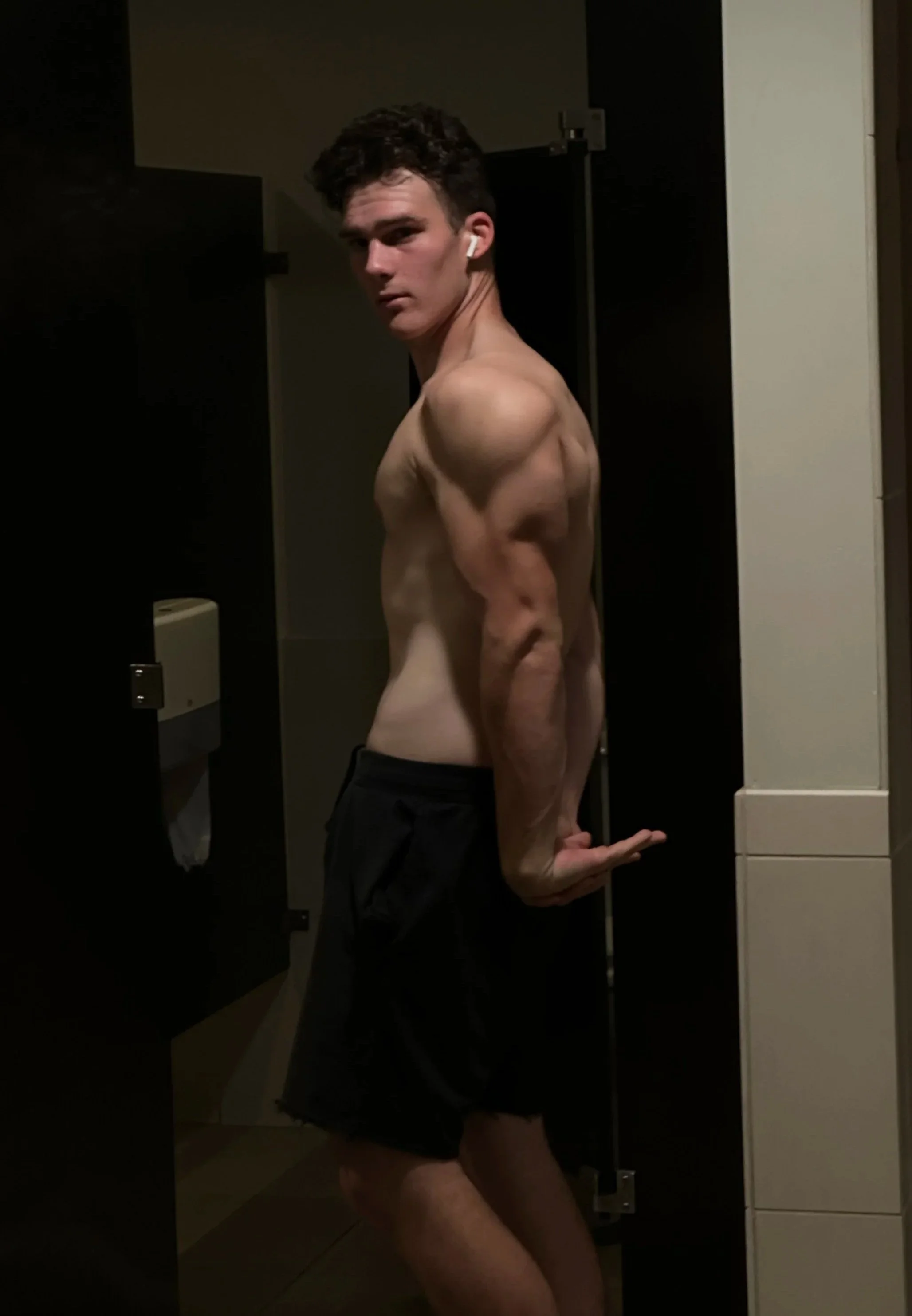 A young man stands shirtless in a bathroom, flexing his right arm, wearing black shorts, with dark curly hair, and AirPods in his ears.