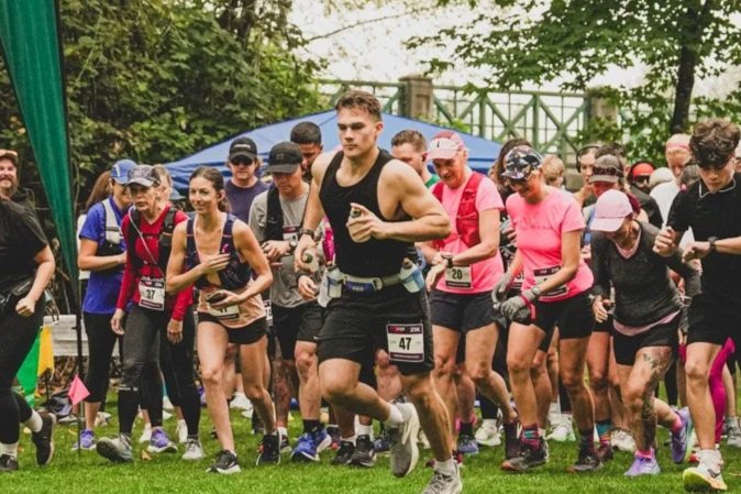 Group of runners starting a race outdoors on grass, with trees and a fence in the background.