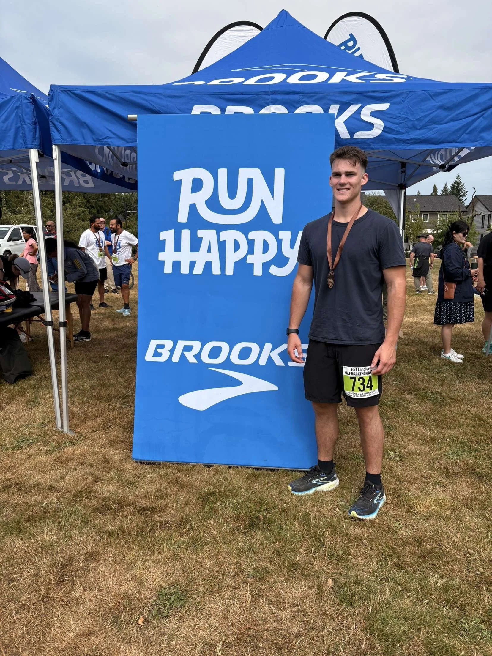 A young man in athletic clothing with a race bib number 734 and a medal around his neck standing next to a large blue banner with white text that reads "RUN HAPPY" and the Brooks logo at a running event.