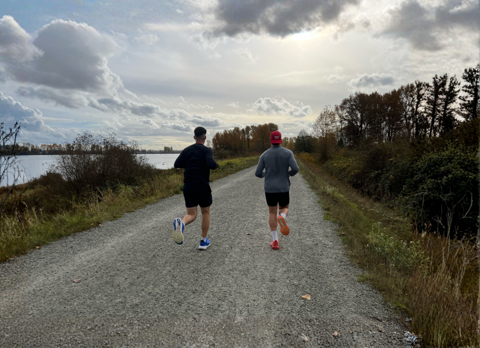 Two people jogging on a gravel trail beside a body of water, with trees and a cloudy sky in the background.