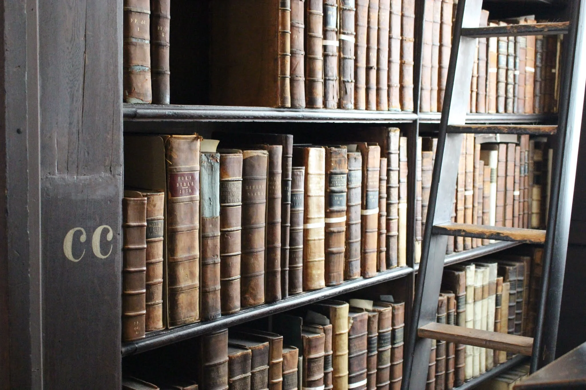 A wooden bookshelf filled with old, worn books in a library, with a ladder leaning against the side.