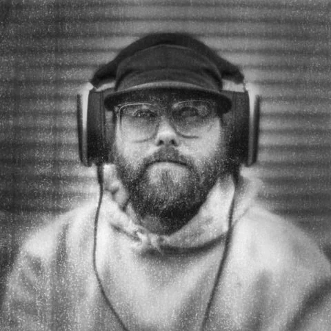 A man with a beard wearing glasses, headphones, a cap, and a hoodie, sitting in front of a textured background.