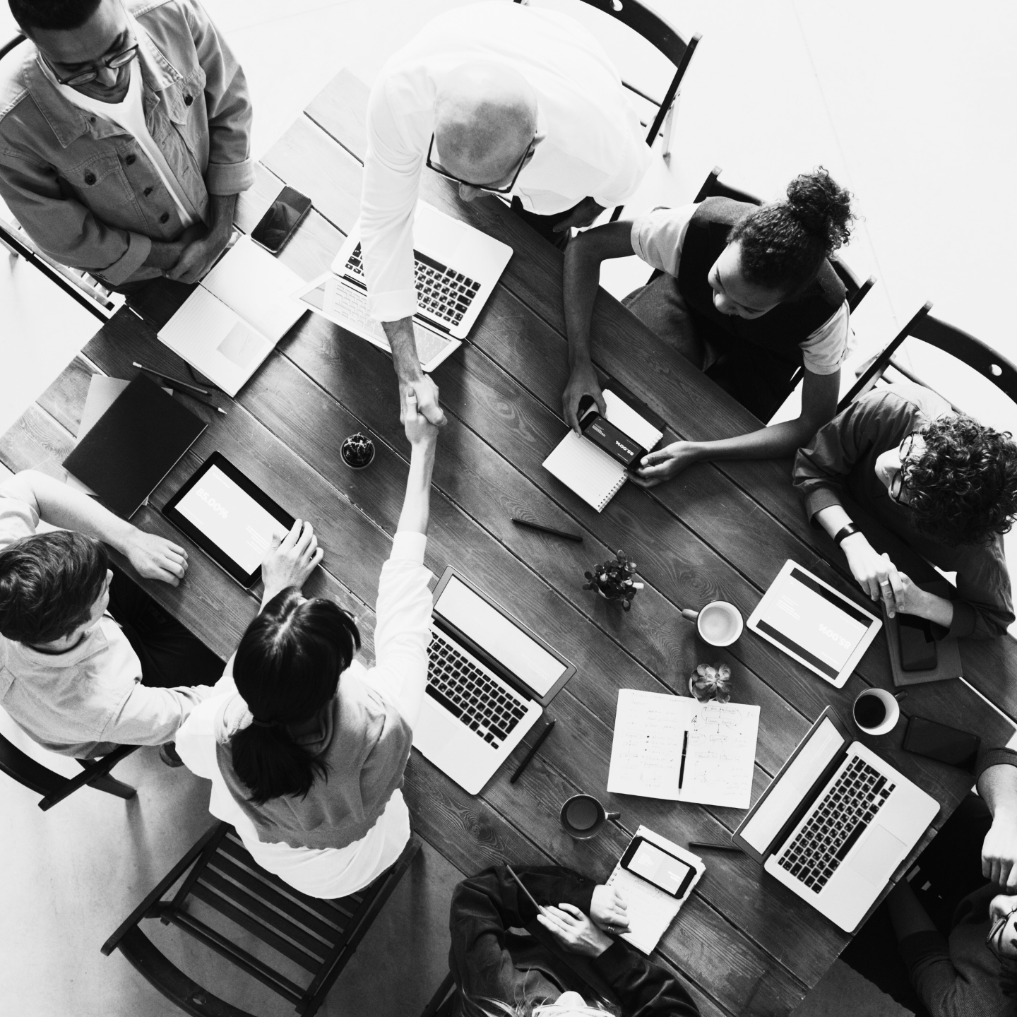 People sitting around a large conference table using laptops and tablets, with two individuals shaking hands across the table.