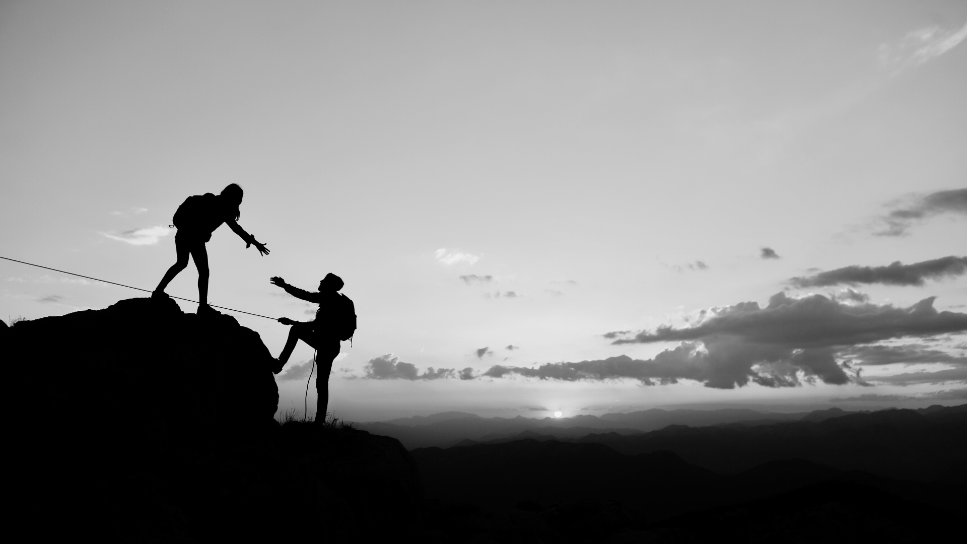 Silhouette of two hikers helping each other climb a mountain at sunset, with a mountain range in the background.