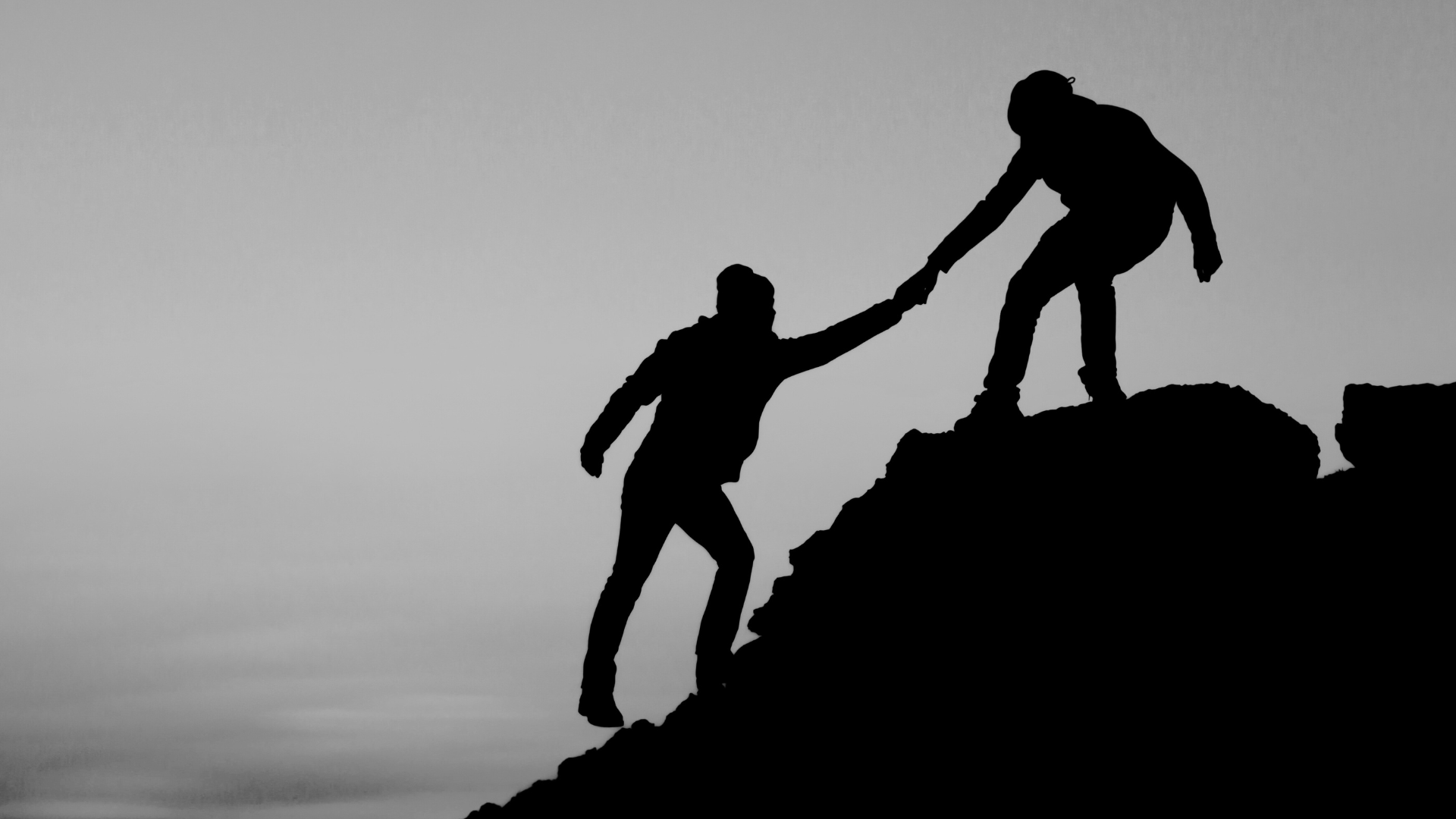 Silhouette of two people on a mountain, one helping the other to climb up.