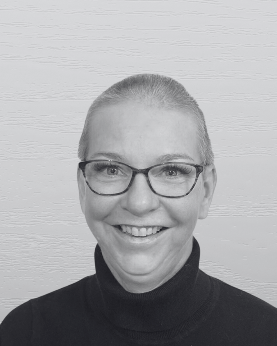 Black and white photo of a smiling woman with short hair and glasses, wearing a turtleneck sweater, standing against a textured wall background.