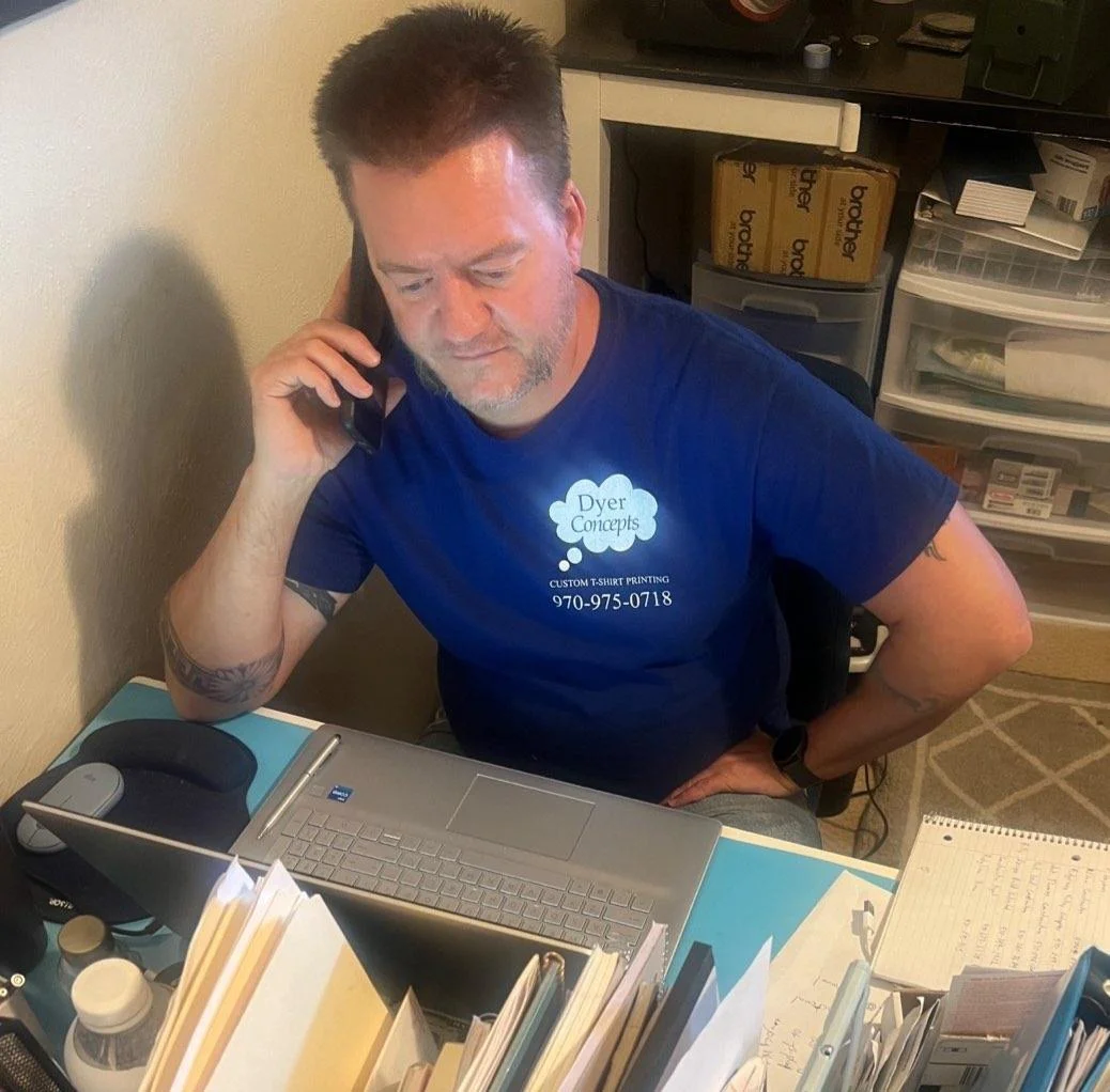 A man sitting at a desk, talking on a phone, with a laptop, mouse, notebook, and various office supplies on the desk. He is wearing a blue t-shirt with the text 'Dyer Concepts' and the phone number 970-975-0718