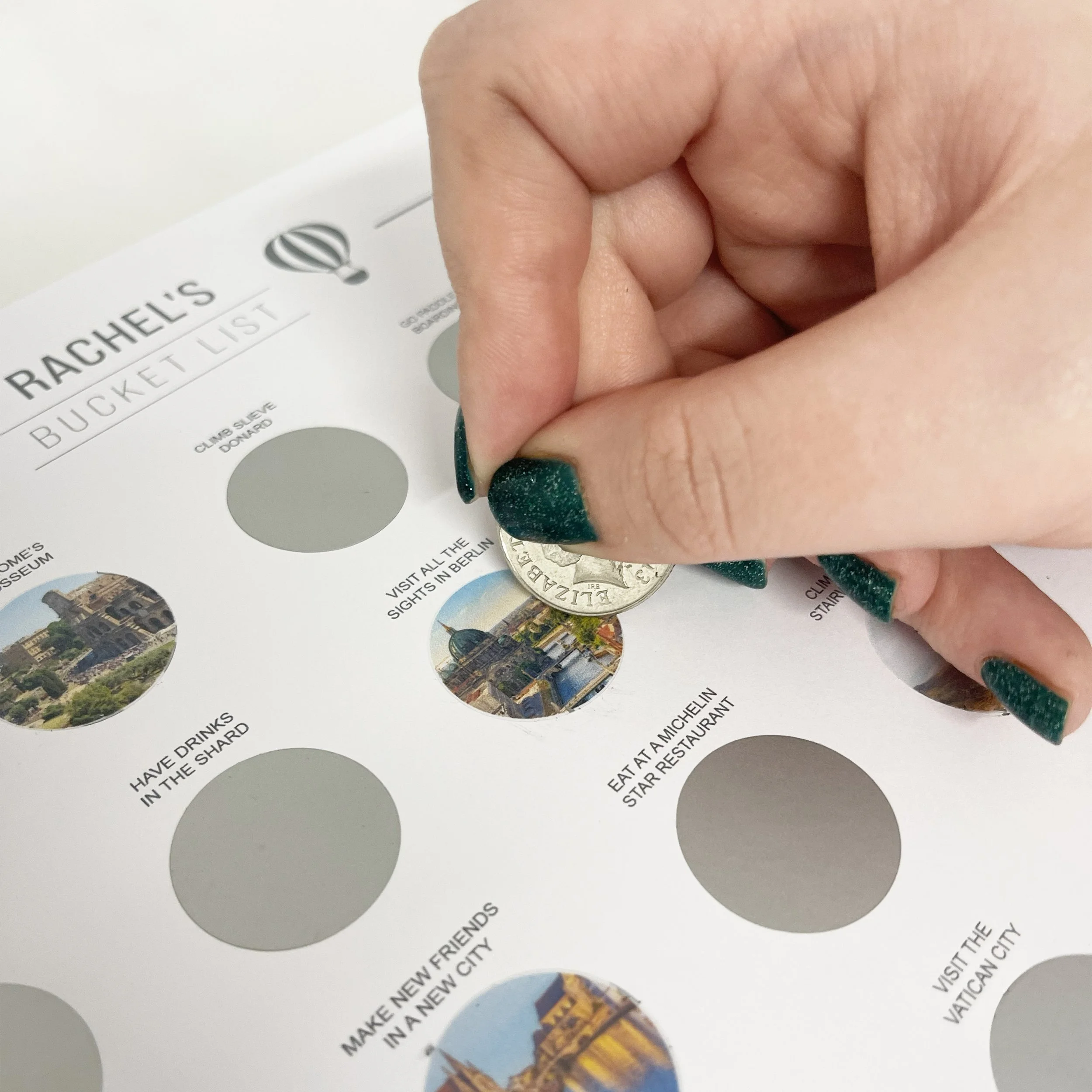 A person using a coin to scratch off a bucket list sheet poster titled 'Rachel's Bucket List', which includes items like visiting the Berlin sights and eating at a Michelin star restaurant.