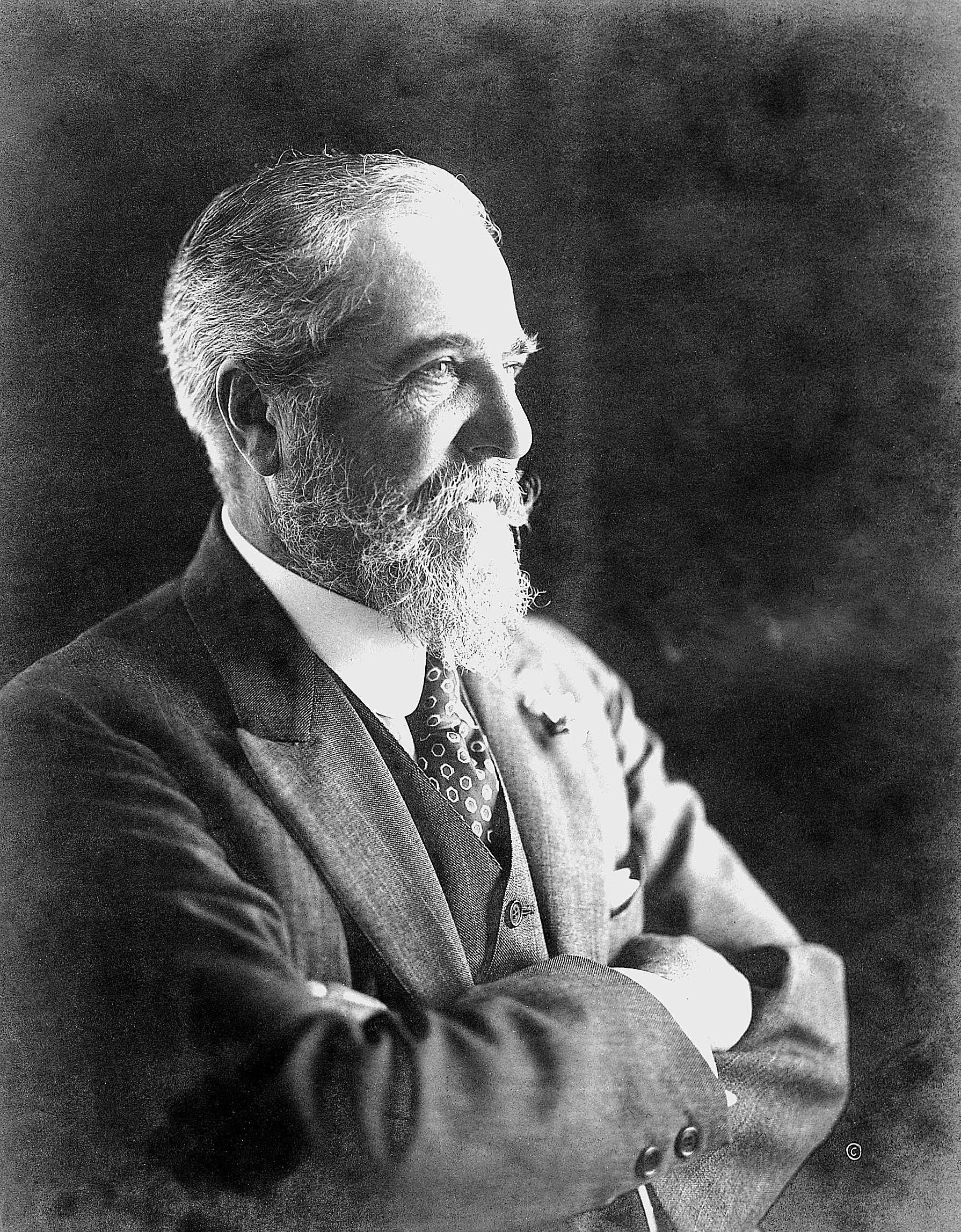 Portrait en noir et blanc d'un homme âgé à barbe et à cheveux gris, habillé en costume, avec ses bras croisés.