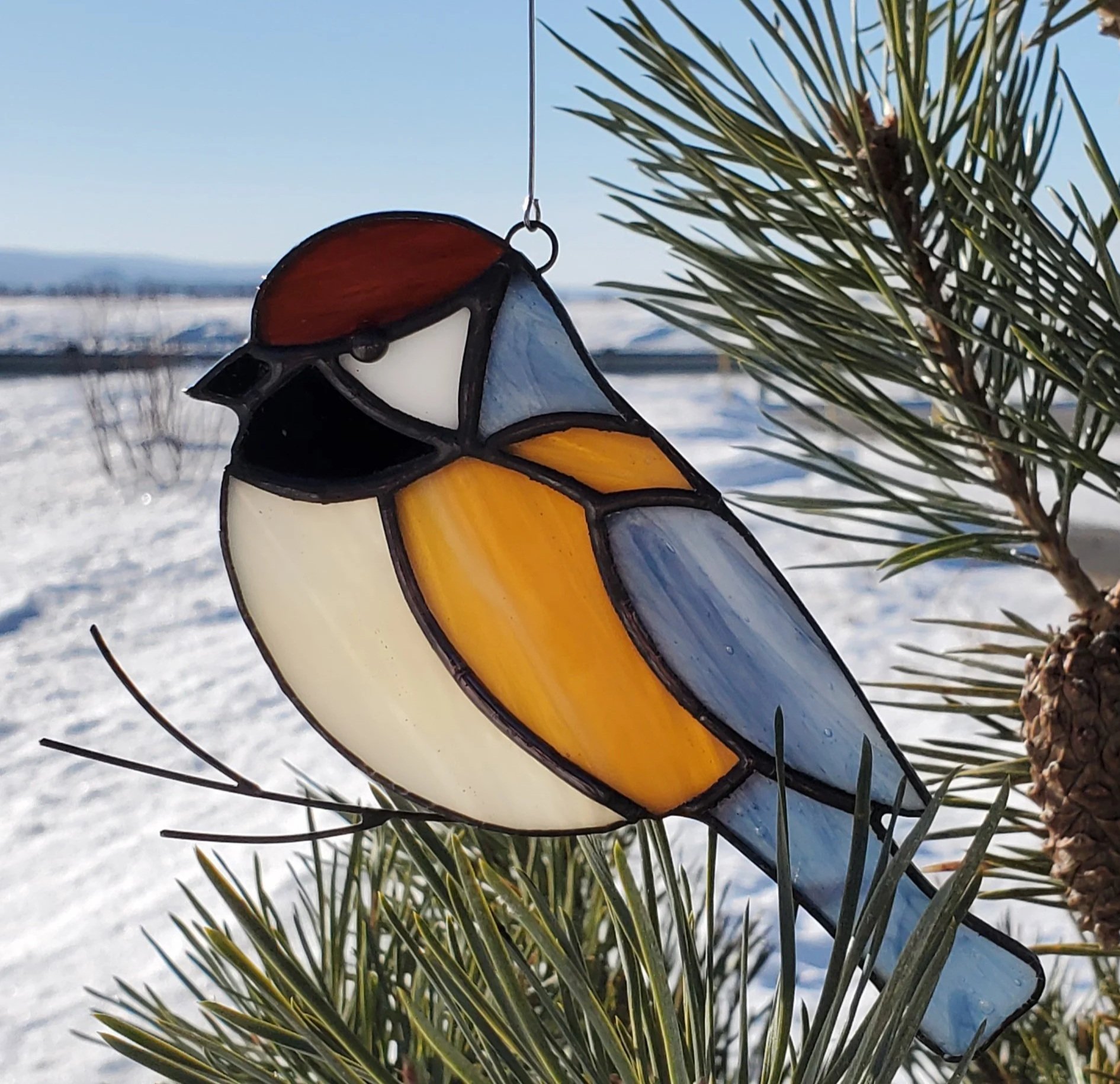 Décoration en verre de stained glass représentant un oiseau coloré, suspendue par un fil, accrochée à une branche de pin en extérieur avec neige et ciel bleu en fond.