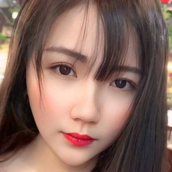 Close-up of a young woman with straight dark hair, fair skin, red lipstick, and makeup, looking directly at the camera.