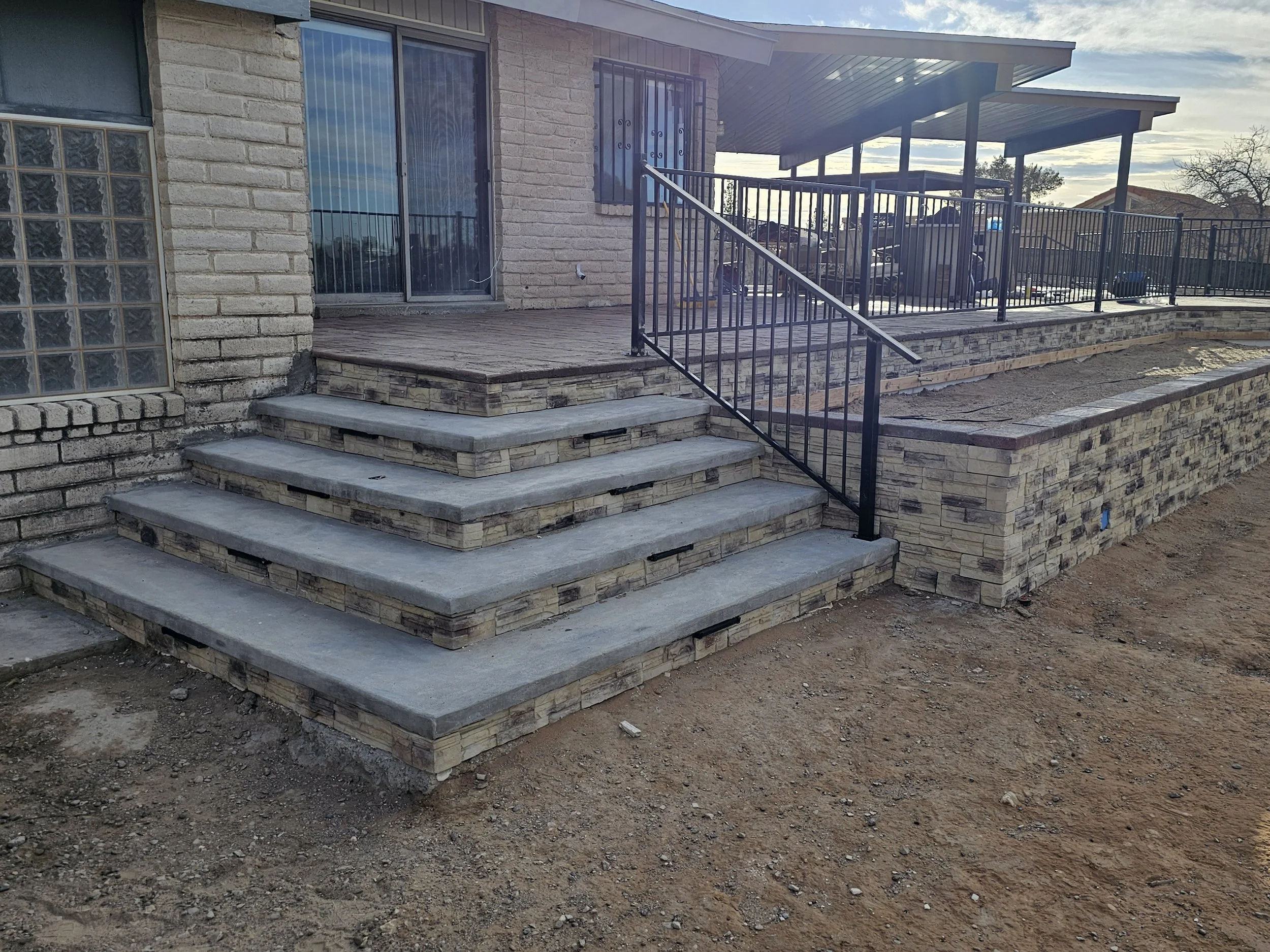 Construction site showing a house with a new stone and concrete staircase leading to a raised patio with a metal railing.