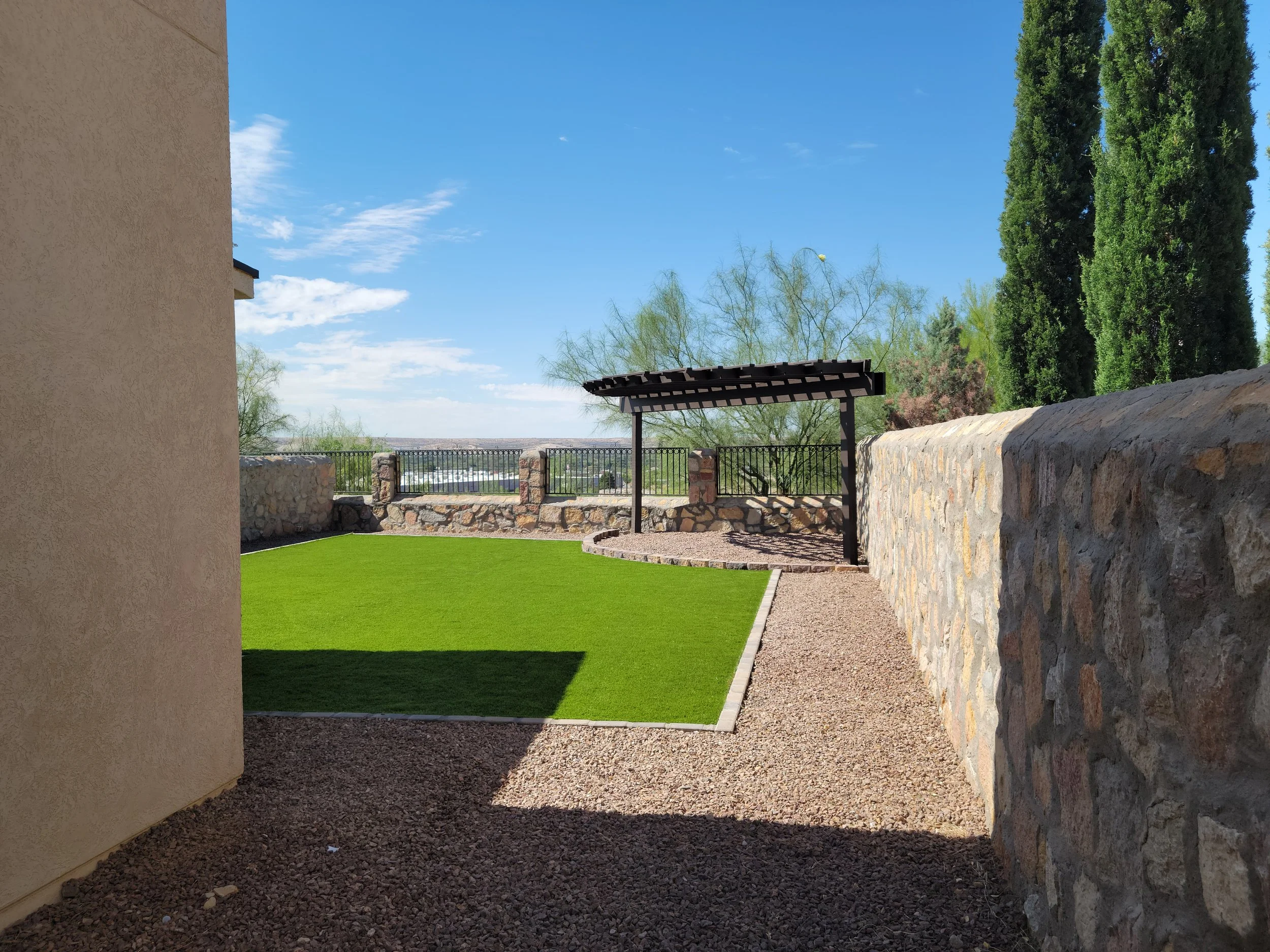 Backyard with a small patch of green artificial grass, a stone wall, and a covered patio area with a metal railing, under a bright blue sky with some clouds.