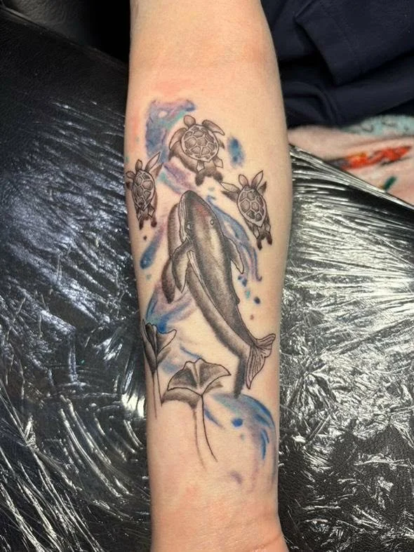 Tattoo of a whale swimming above water, surrounded by three turtles and two stingrays, with blue watercolor-style splashes in the background.