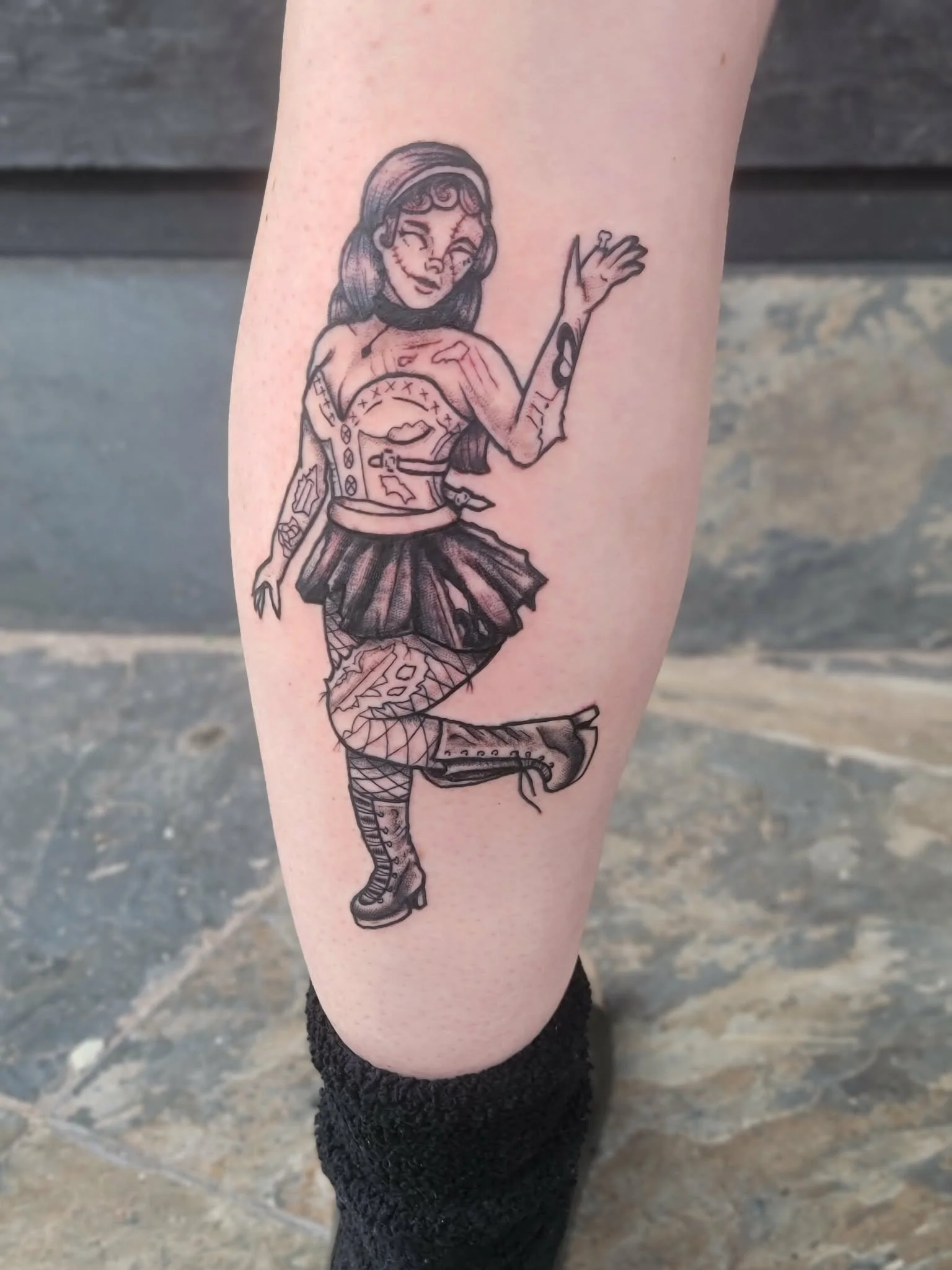Black and gray tattoo of a woman with tattoos in a punk style, wearing fishnet stockings, a skirt, and combat boots, with one arm raised.