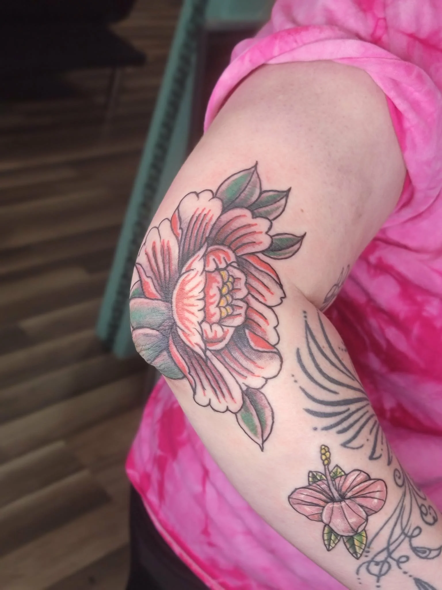 Tattoo of a large flower with leaves on an arm, and a smaller flower on lower arm, with a pink tie-dye shirt.