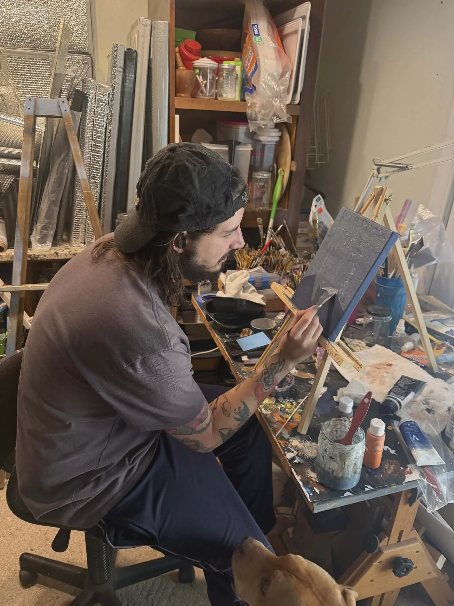 A man with long hair, tattoos, wearing a black cap backwards and a gray T-shirt, is sitting on a chair in an art studio, painting on a canvas set on an easel. A golden retriever is looking up at him. The workspace is cluttered with various art supplies, tools, and miscellaneous items.