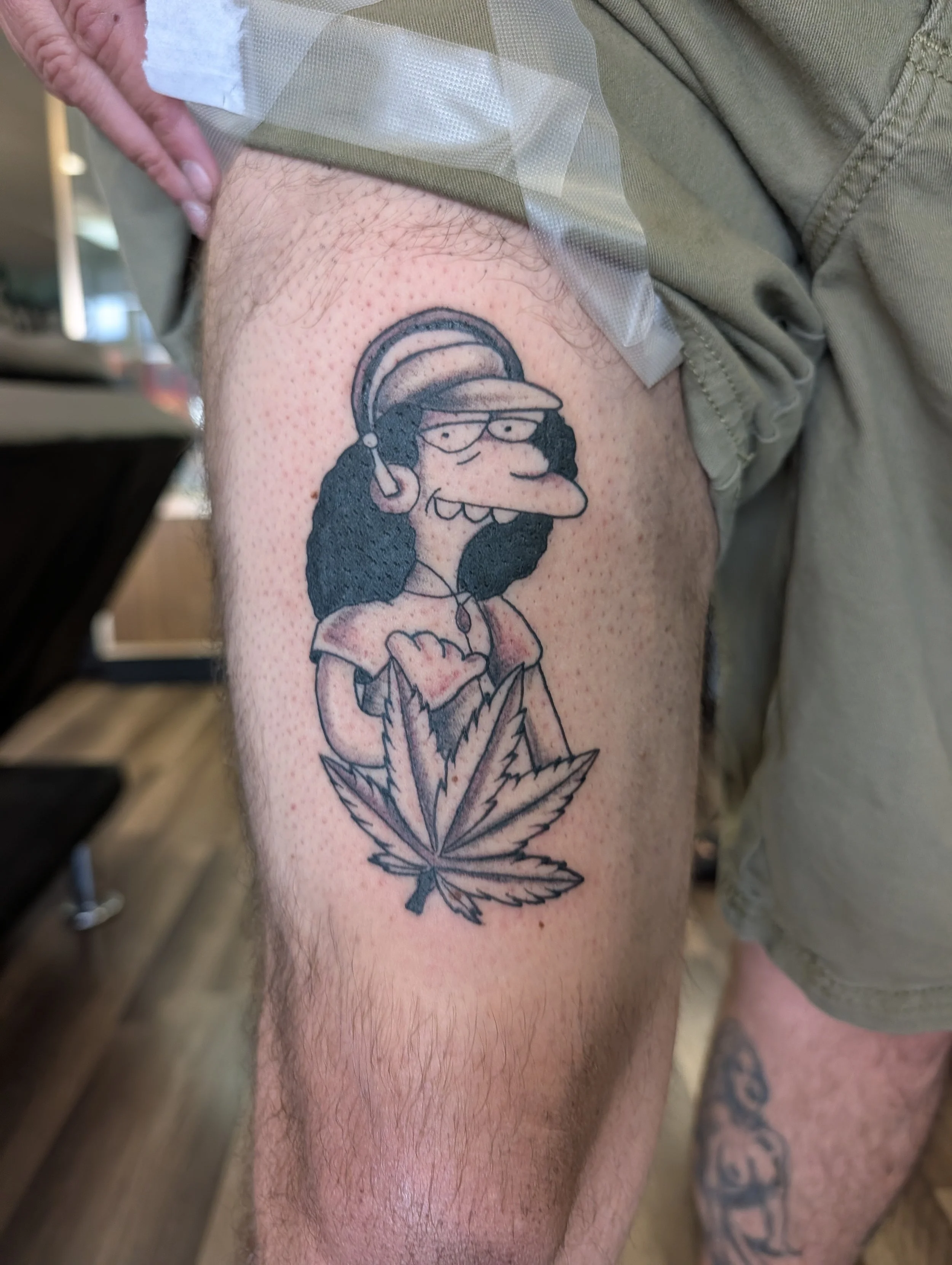 Tattoo of a cartoon character with glasses, a cap, headphones, and a necklace, sitting on a marijuana leaf.
