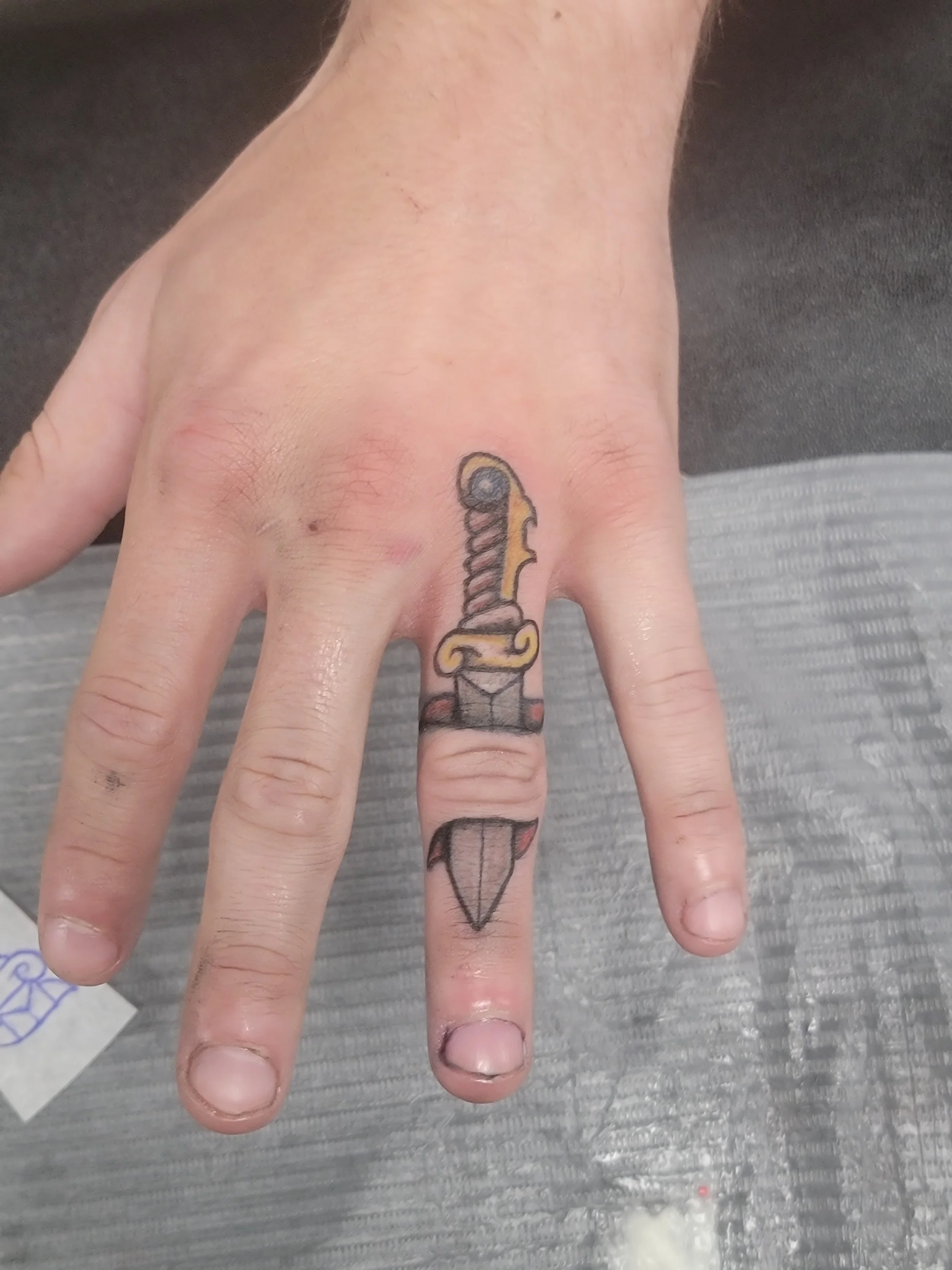 A hand with a colorful tattoo of a sword on the middle finger, resting on a gray surface. The tattoo features a yellow and black hilt with a red base.