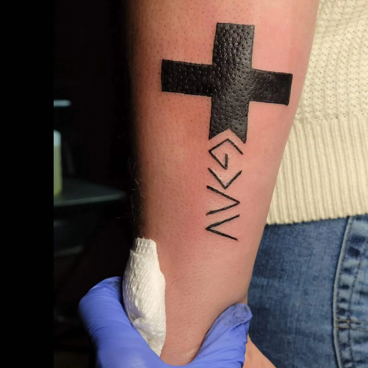 Tattoo of a black cross  on a person's forearm, with a person wearing a blue glove holding around the wrist.