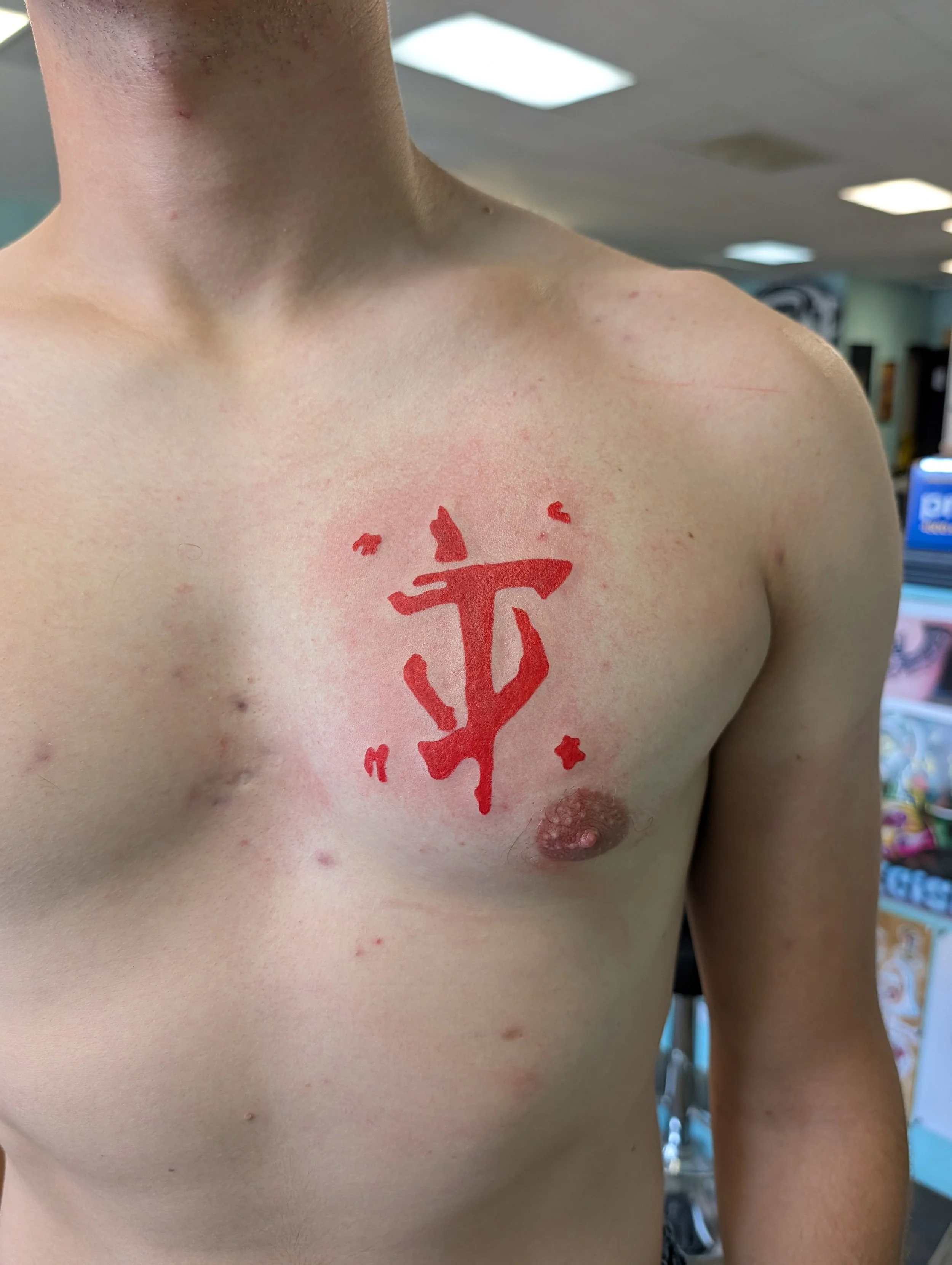 A close-up of a person's chest showing a red tattoo of a dreamcatcher with a cross and stars, with the person's skin showing some freckles and small spots.