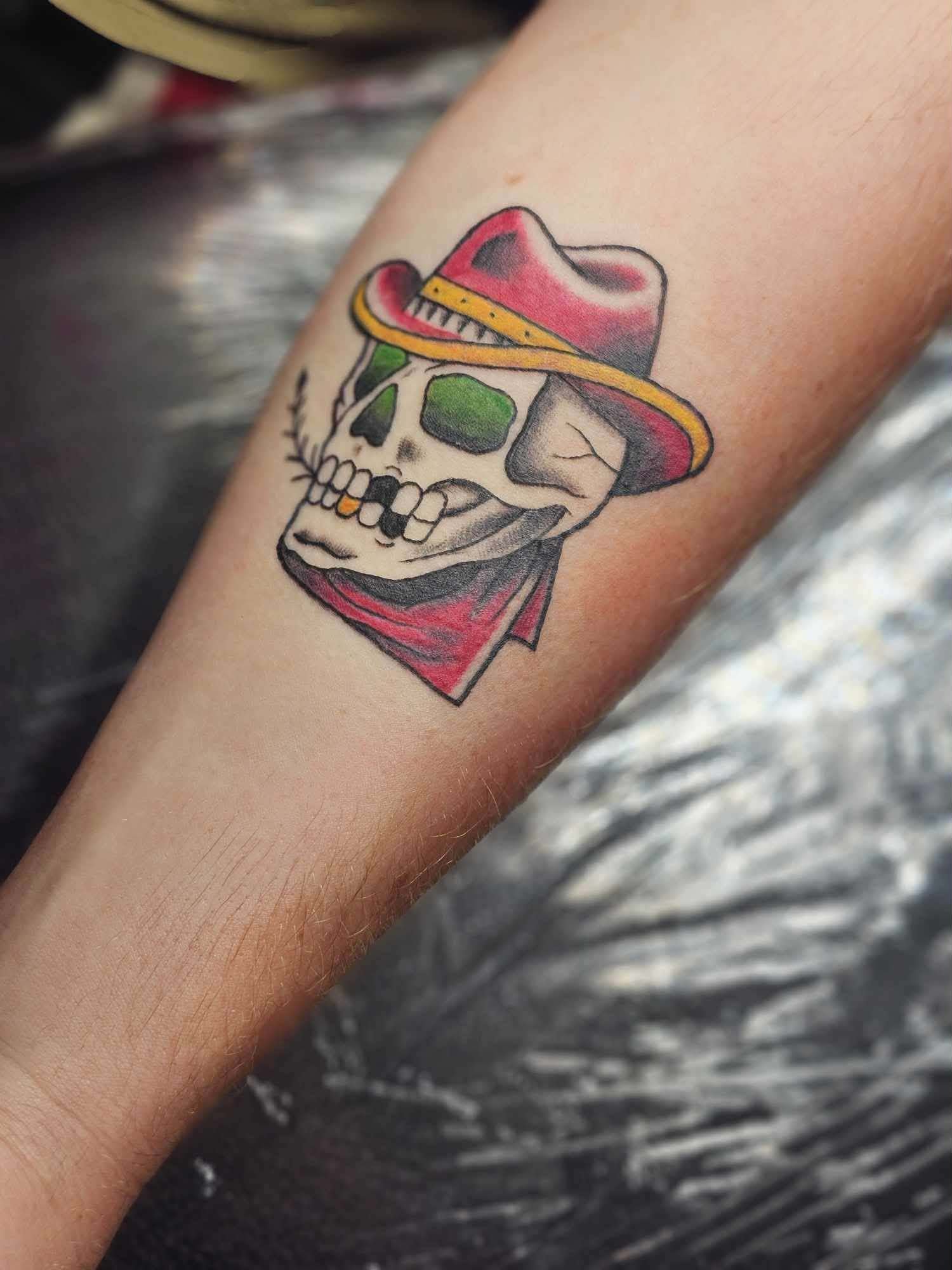 Tattoo of a skull with a green eye socket, wearing a red bandana, and a pink and black cowboy hat with a yellow band, on a person's forearm in traditional style.