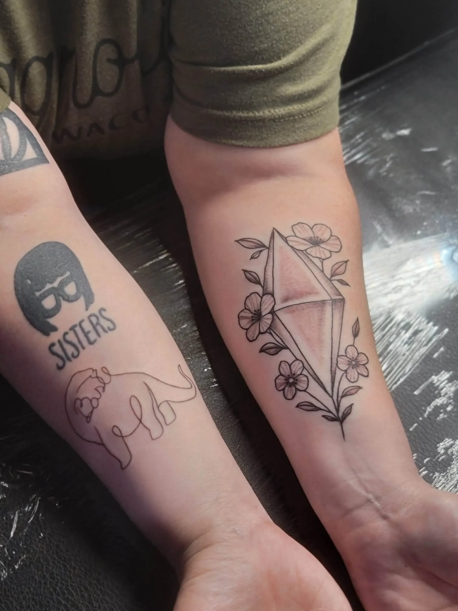 Two forearms with tattoos: one features a black skull with glasses and the word 'SISTERS' underneath, the other has a geometric diamond surrounded by flowers.