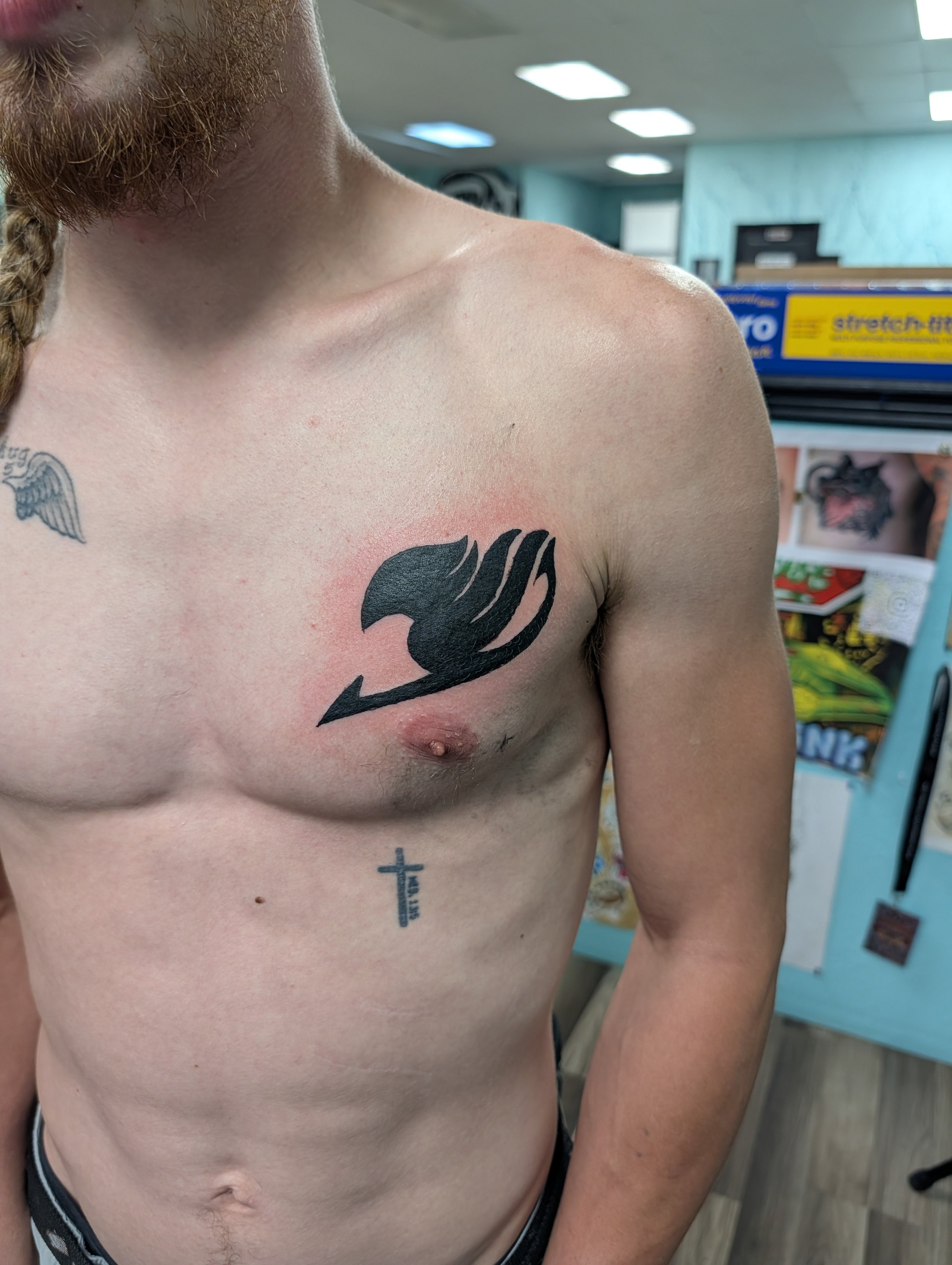 A close-up of a person's upper torso showing tattoos on the chest and side, with a tattoo of a whale and a cross with text, in a room with bright fluorescent lighting and shelving units in the background.